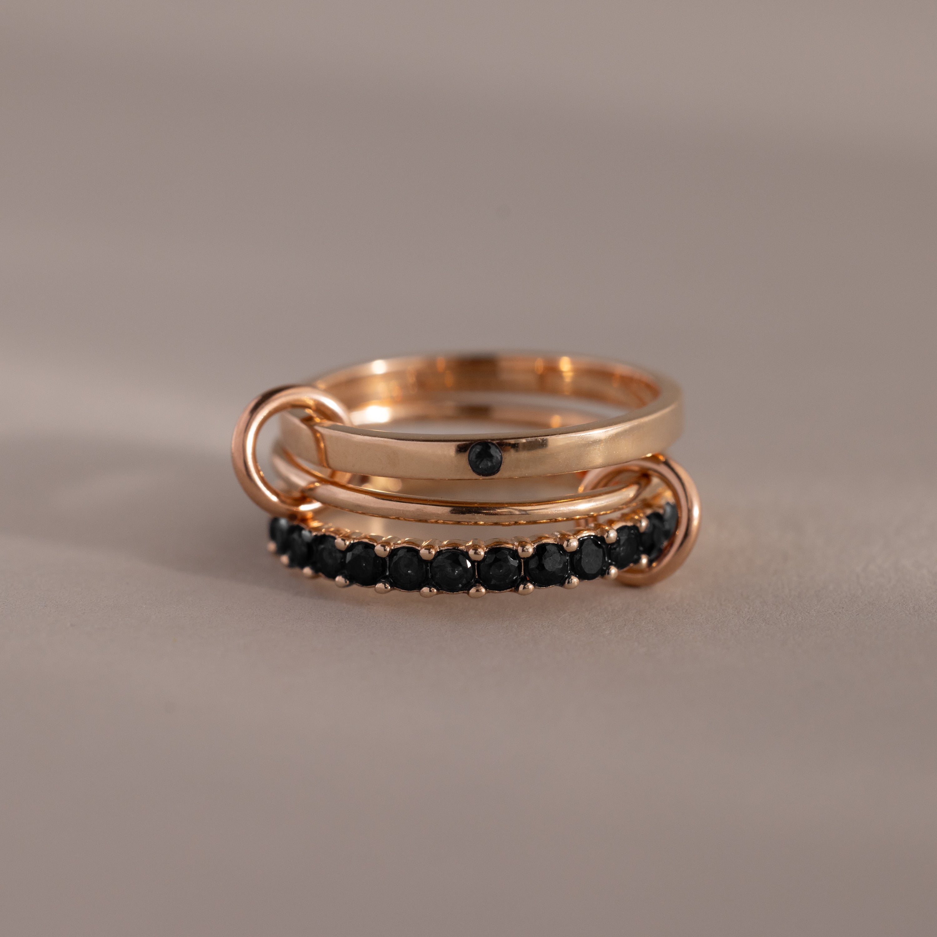 The Triple Linked Onyx Ring Set features rose gold rings linked together—one plain with a black stone and the other lined with small black stones—offering an elegant, stackable, modern birthstone band.