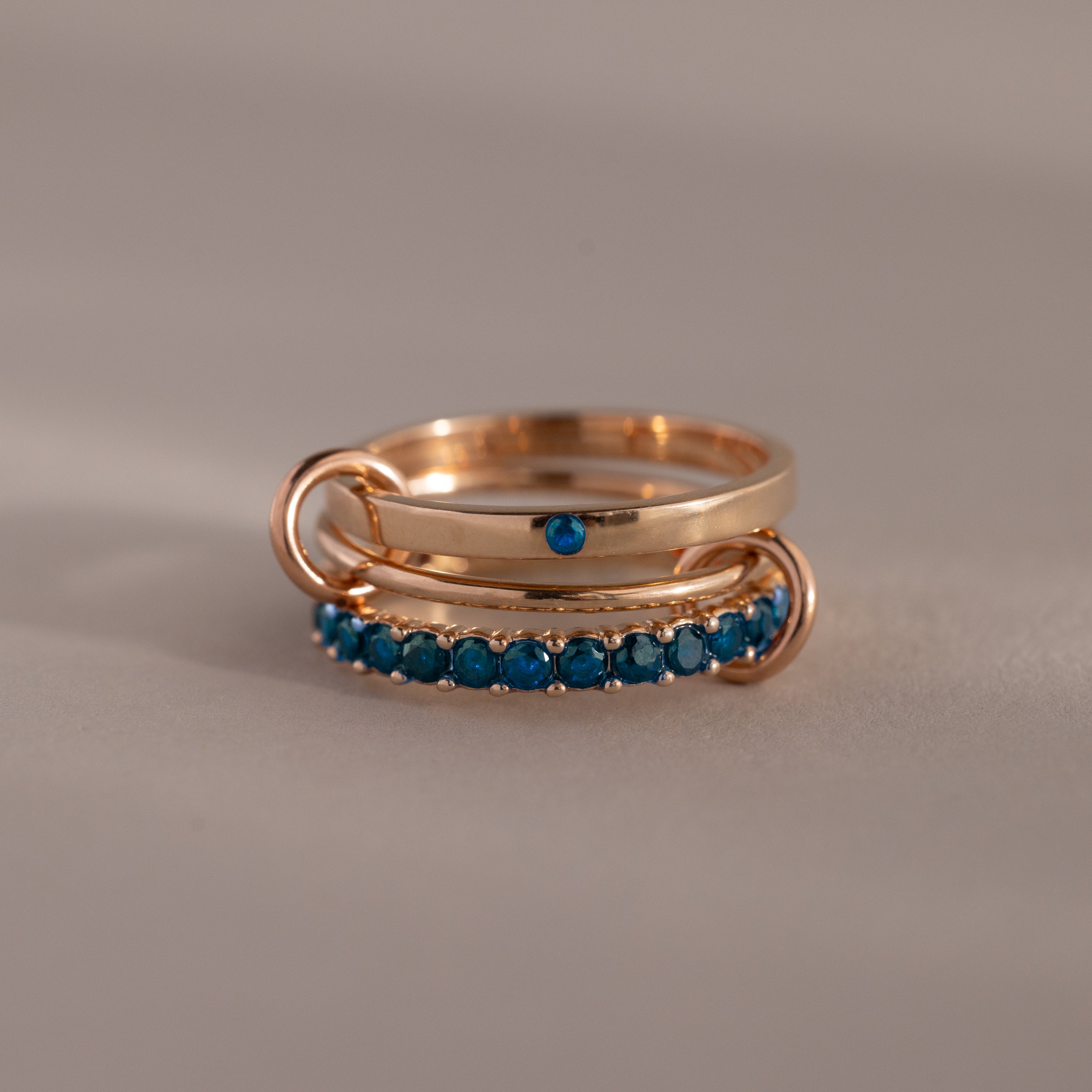 The Triple Linked Blue Zircon Ring Set features rose gold bands with rows of small blue zircons and a single blue stone, shown on a neutral background.