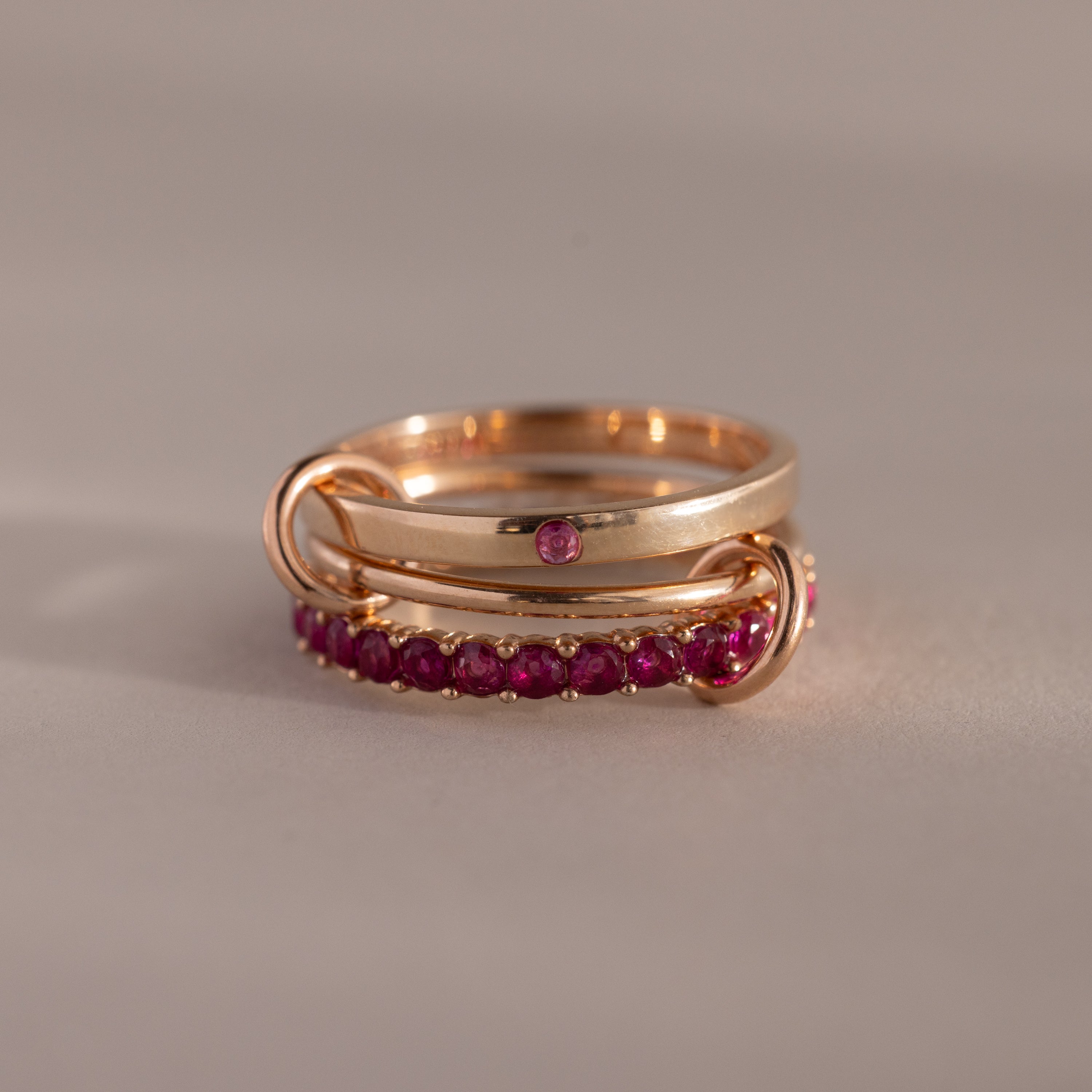 The Triple Linked Ruby Ring Set features three gold rings—one plain and two adorned with small pink ruby gemstones—elegantly interlinked on a neutral background.