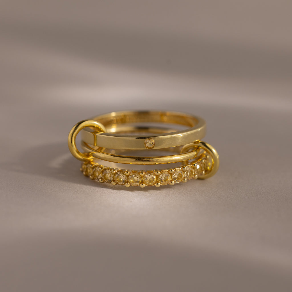 The Triple Linked Topaz Ring Set, featuring three stacked gold bands with delicate topaz accents, is displayed on a neutral background with soft lighting.