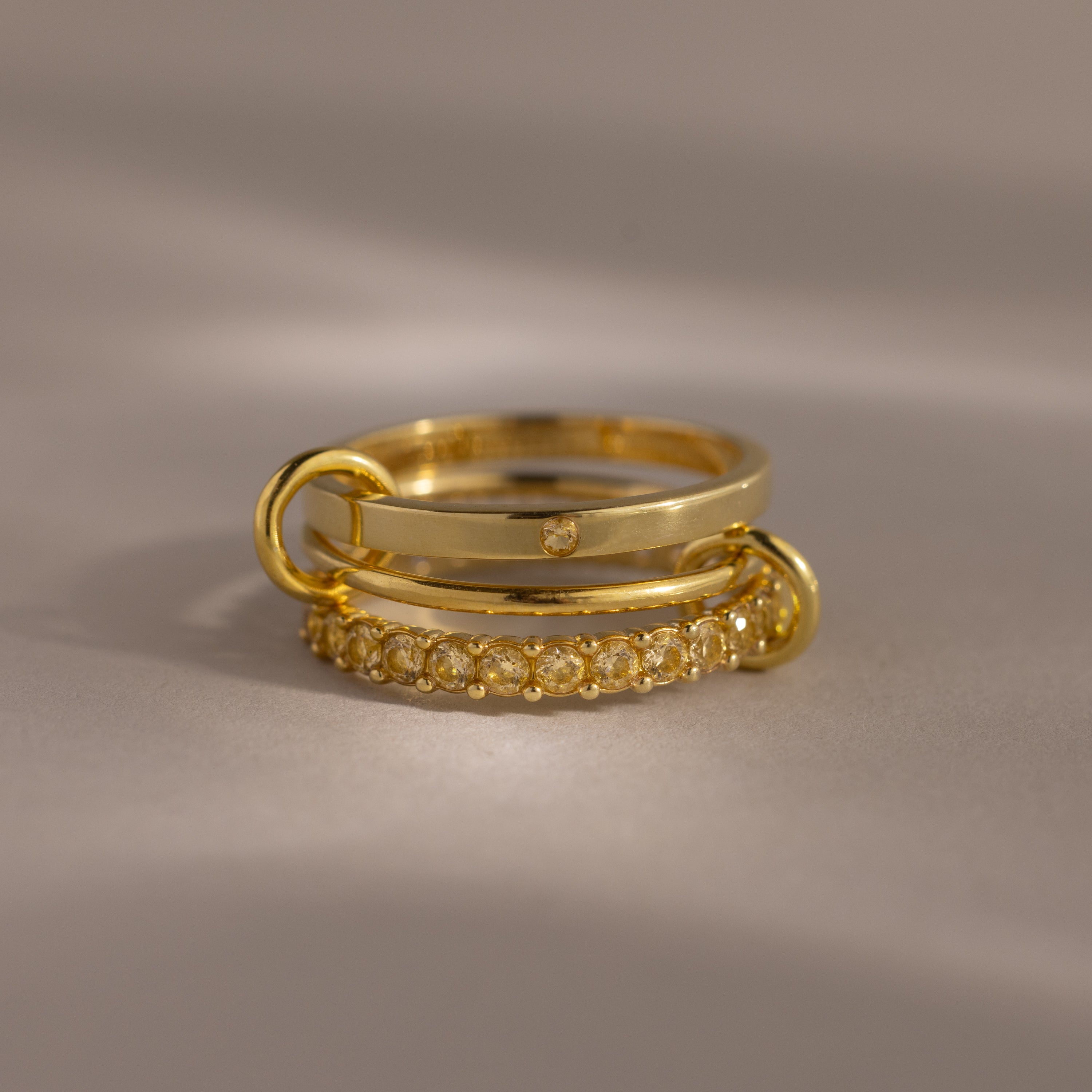 The Triple Linked Topaz Ring Set, featuring three stacked gold bands with delicate topaz accents, is displayed on a neutral background with soft lighting.