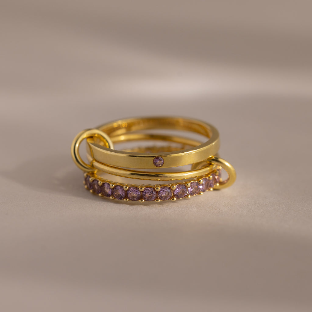The Triple Linked Birthstone Ring Set in Alexandrite (June) features three gold rings, one accented with purple stones, stacked on a neutral background—a gorgeous set for any occasion.