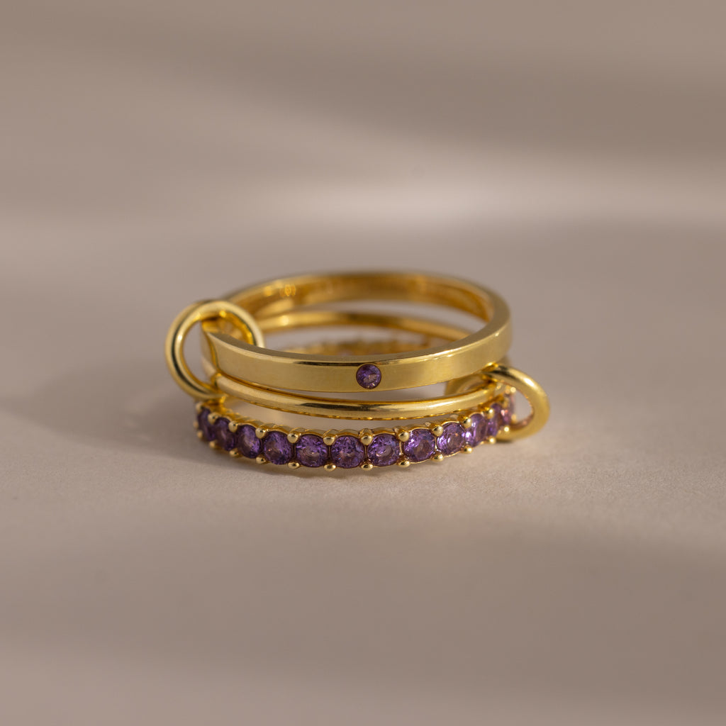 The Triple Linked Amethyst Ring Set features gold rings with purple gemstones, including a stunning birthstone ring and an elegant eternity band, stacked together on a light surface.
