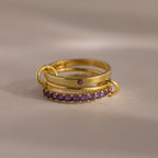 The Triple Linked Amethyst Ring Set features gold rings with purple gemstones, including a stunning birthstone ring and an elegant eternity band, stacked together on a light surface.