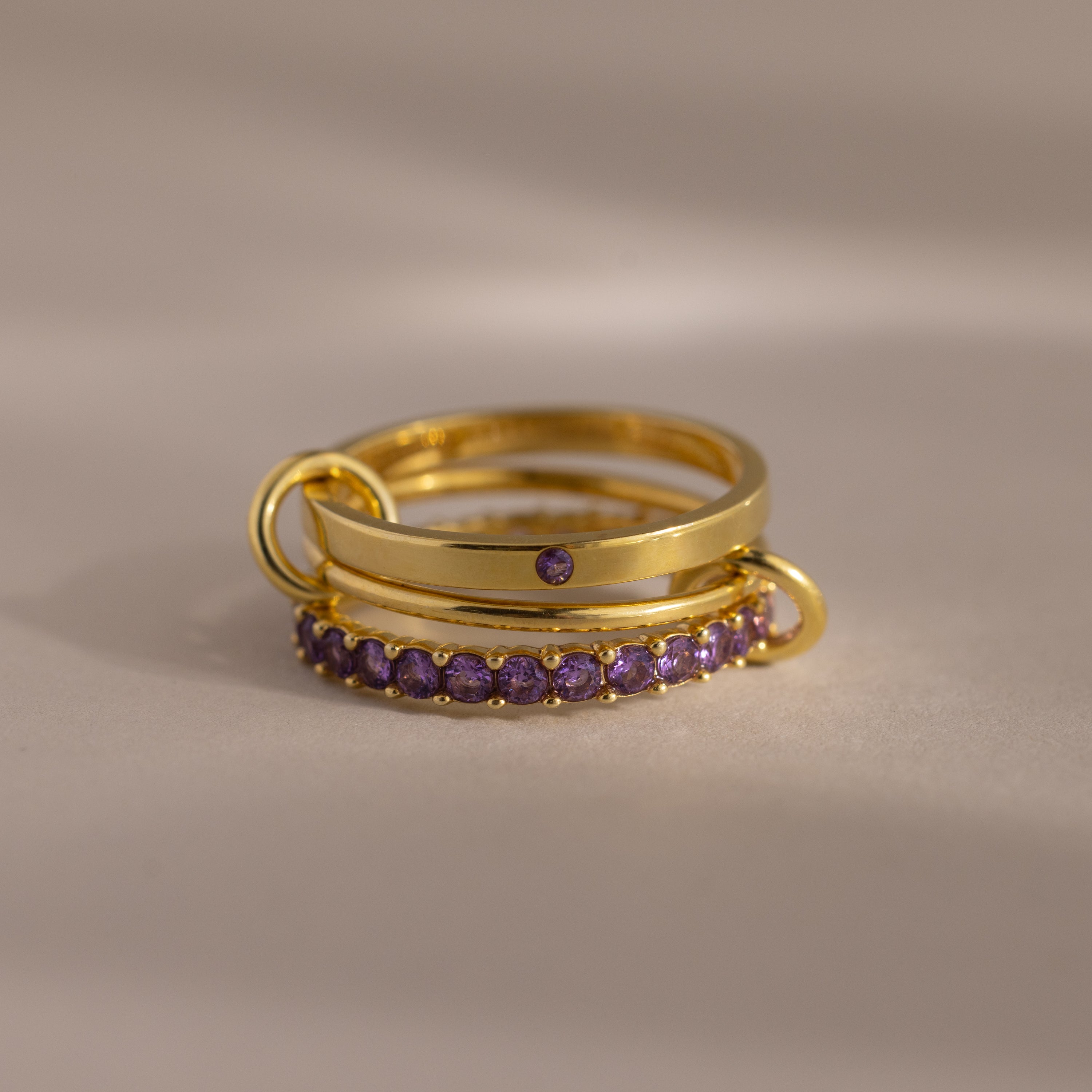 The Triple Linked Amethyst Ring Set features gold rings with purple gemstones, including a stunning birthstone ring and an elegant eternity band, stacked together on a light surface.