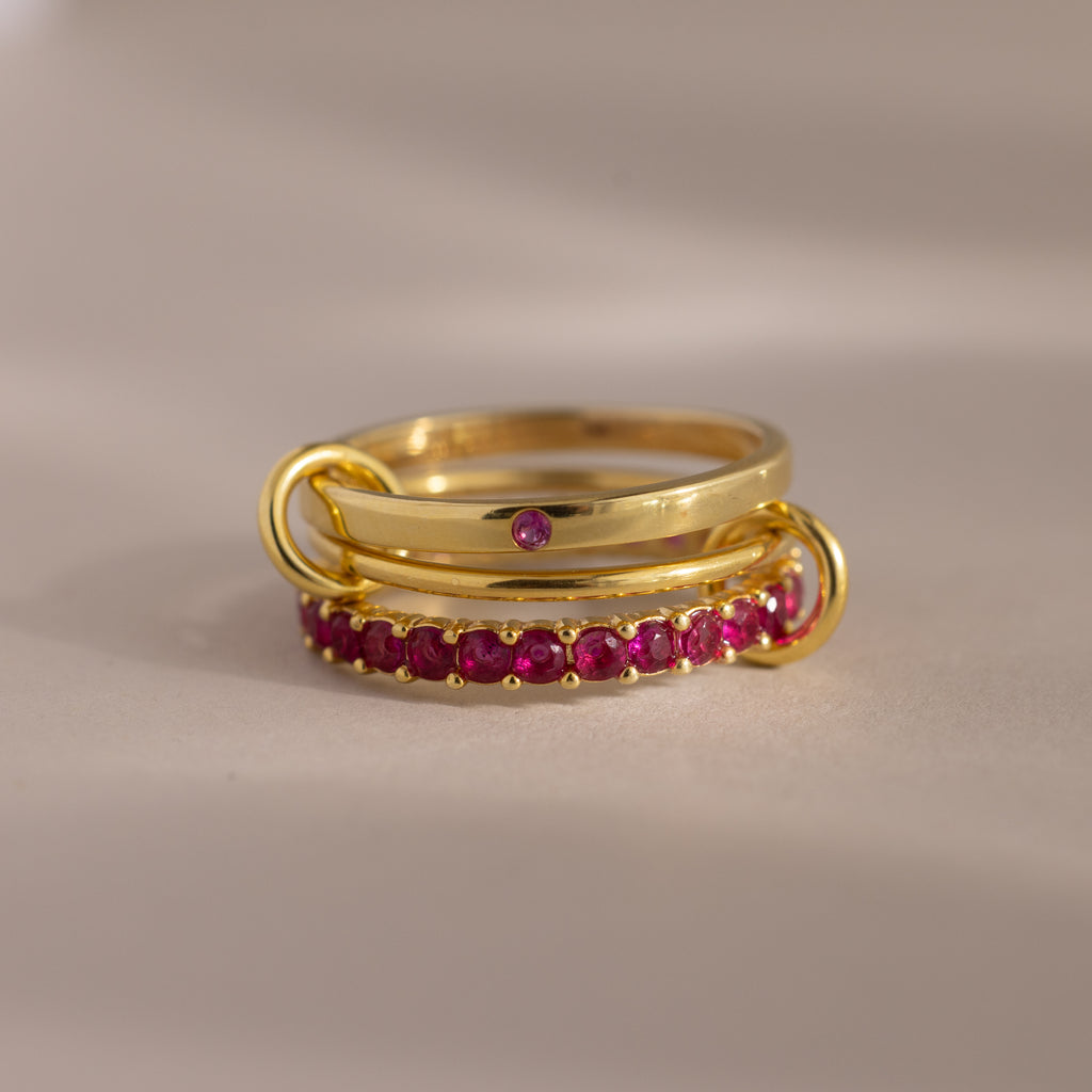 The Triple Linked Ruby Ring Set features three gracefully connected gold rings: one plain with a small ruby and two eternity bands adorned with sparkling rubies.