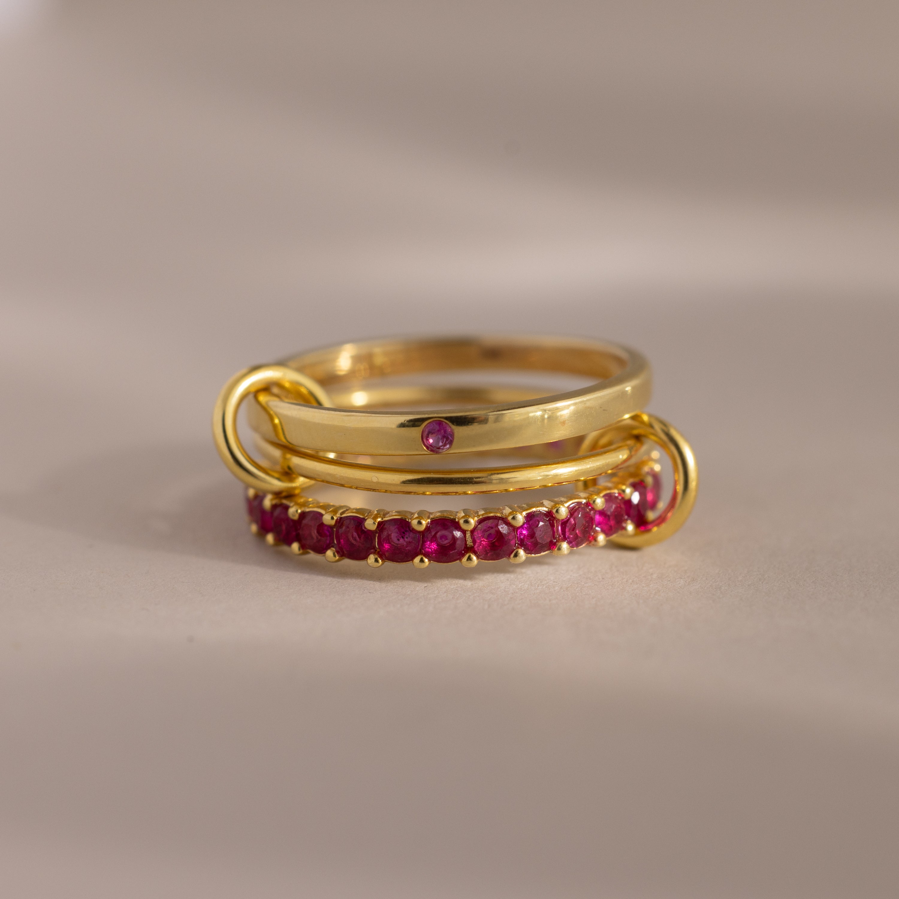 The Triple Linked Ruby Ring Set features three gracefully connected gold rings: one plain with a small ruby and two eternity bands adorned with sparkling rubies.