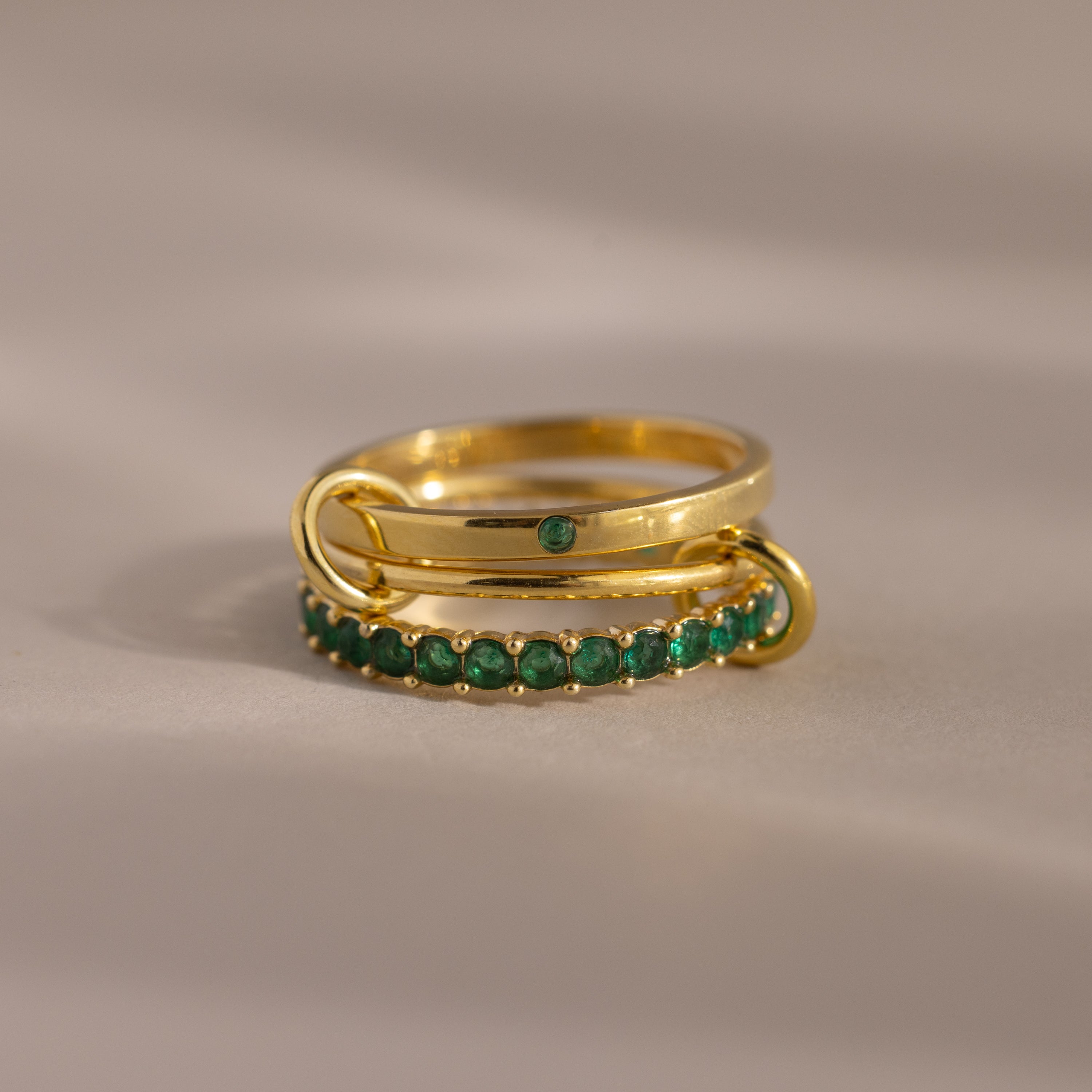 The Triple Linked Emerald Ring Set features two stacked gold rings: an eternity band with green stones and a statement ring with a single green stone, beautifully displayed on a neutral background.
