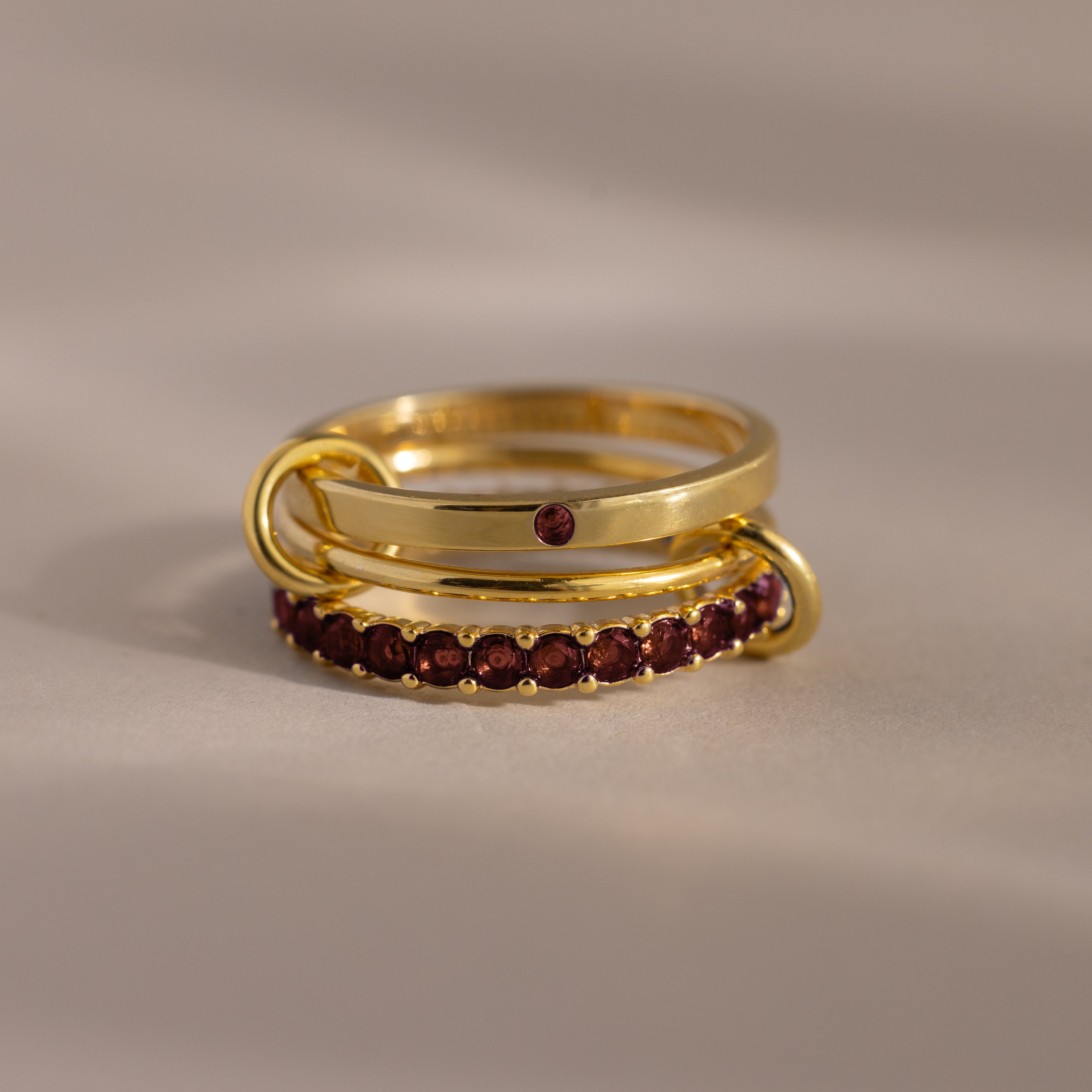 The Triple Linked Garnet Ring Set, featuring gold rings with red gemstones, is stacked together on a neutral surface under soft lighting.
