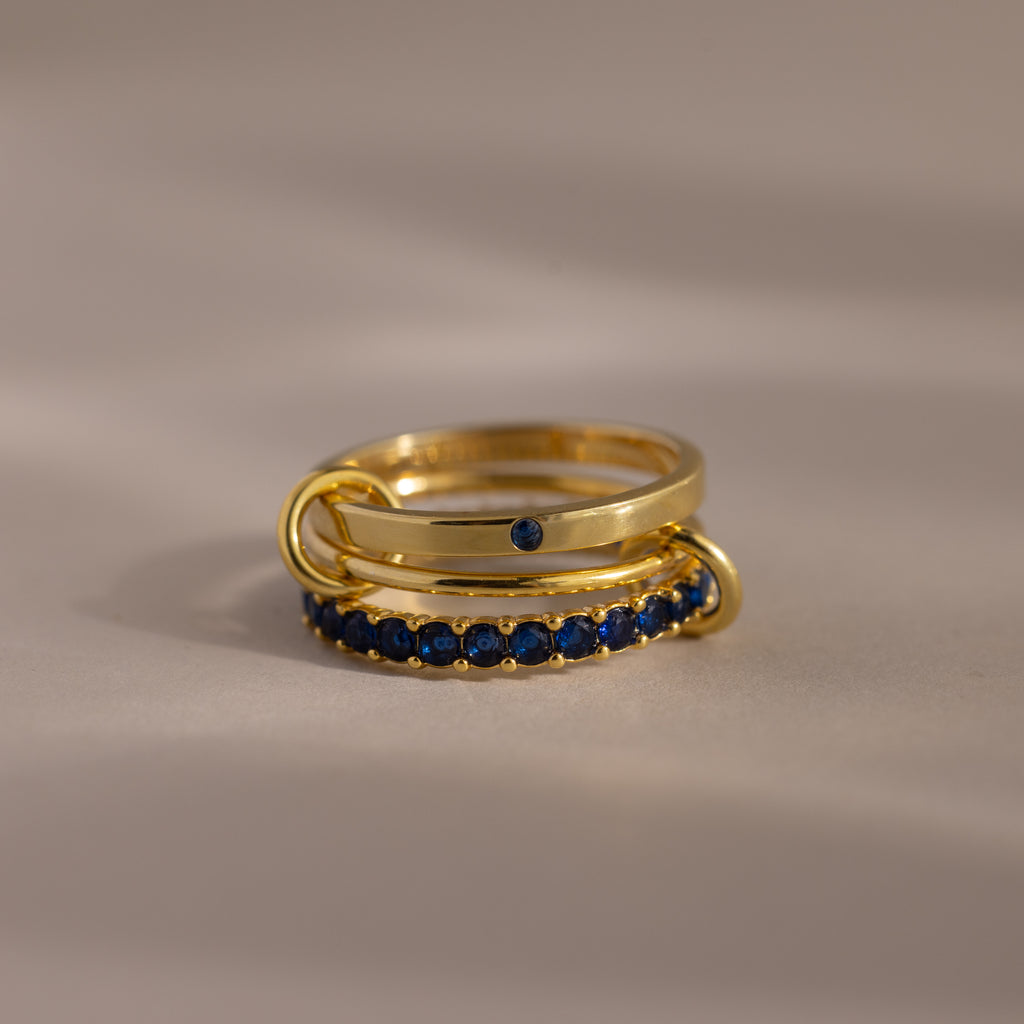 The Triple Linked Sapphire Ring Set, featuring three gold bands adorned with blue sapphires, is displayed on a neutral surface.