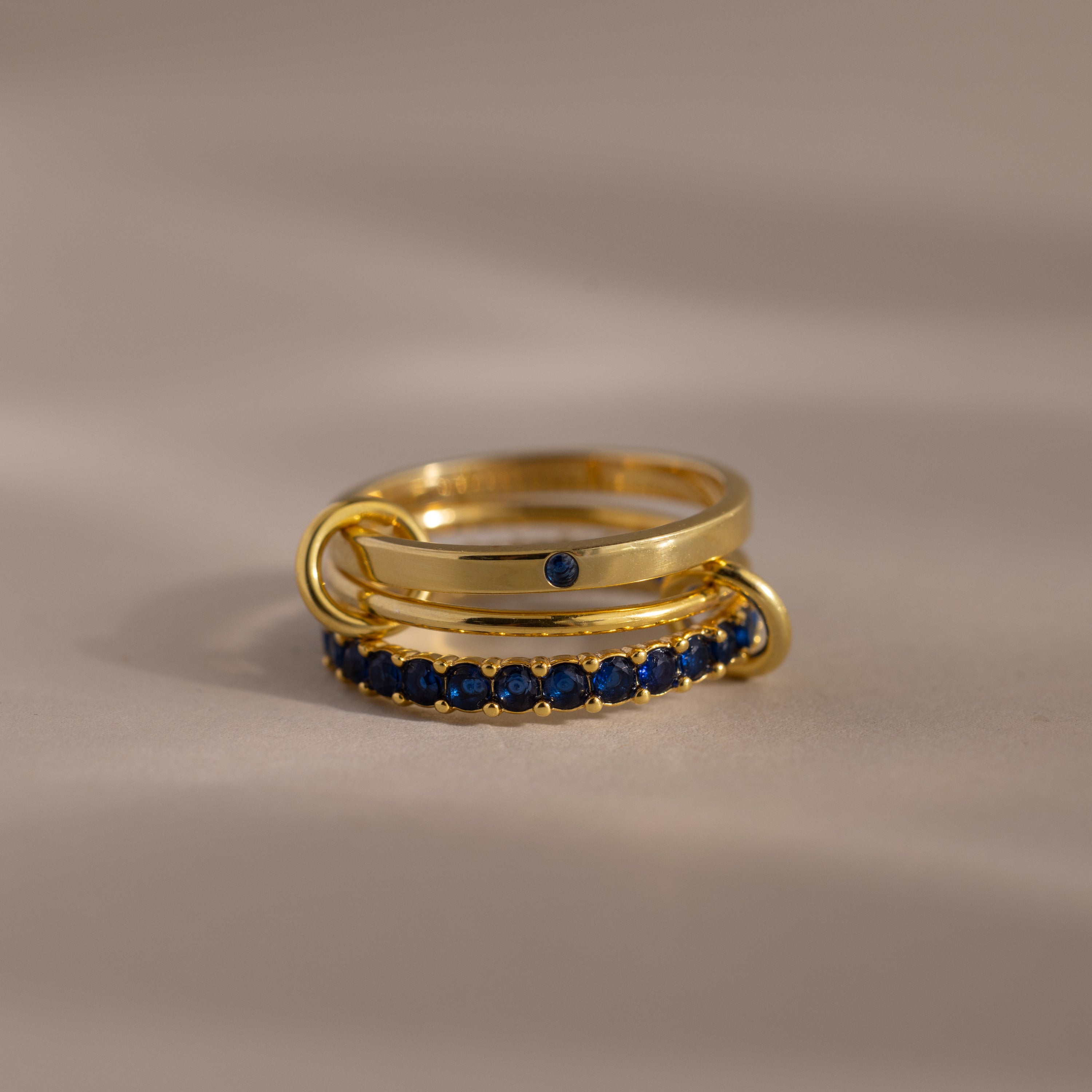 The Triple Linked Sapphire Ring Set, featuring three gold bands adorned with blue sapphires, is displayed on a neutral surface.
