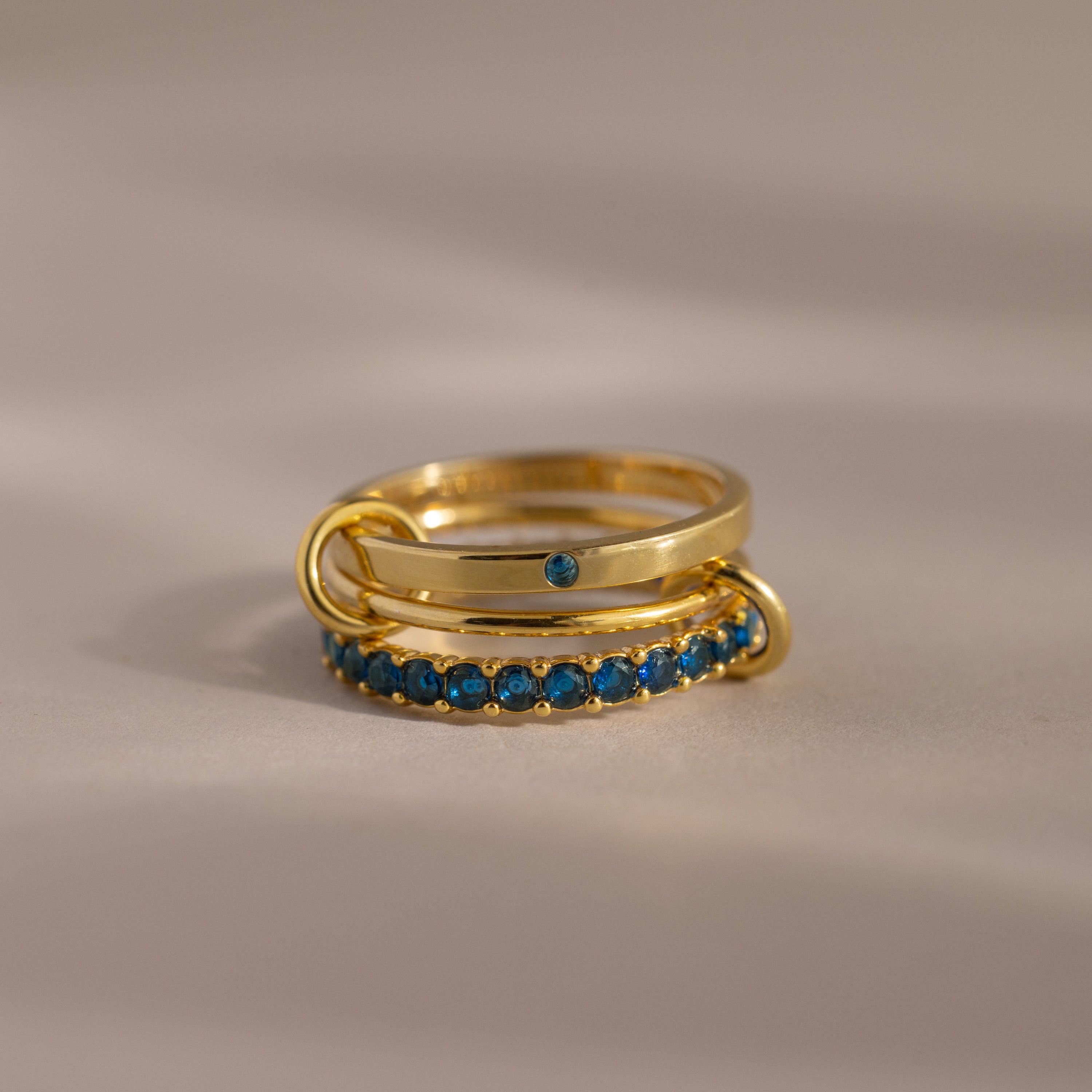 The Triple Linked Blue Zircon Ring Set, featuring gold bands with blue zircon stones, is stacked together on a neutral surface under soft lighting.