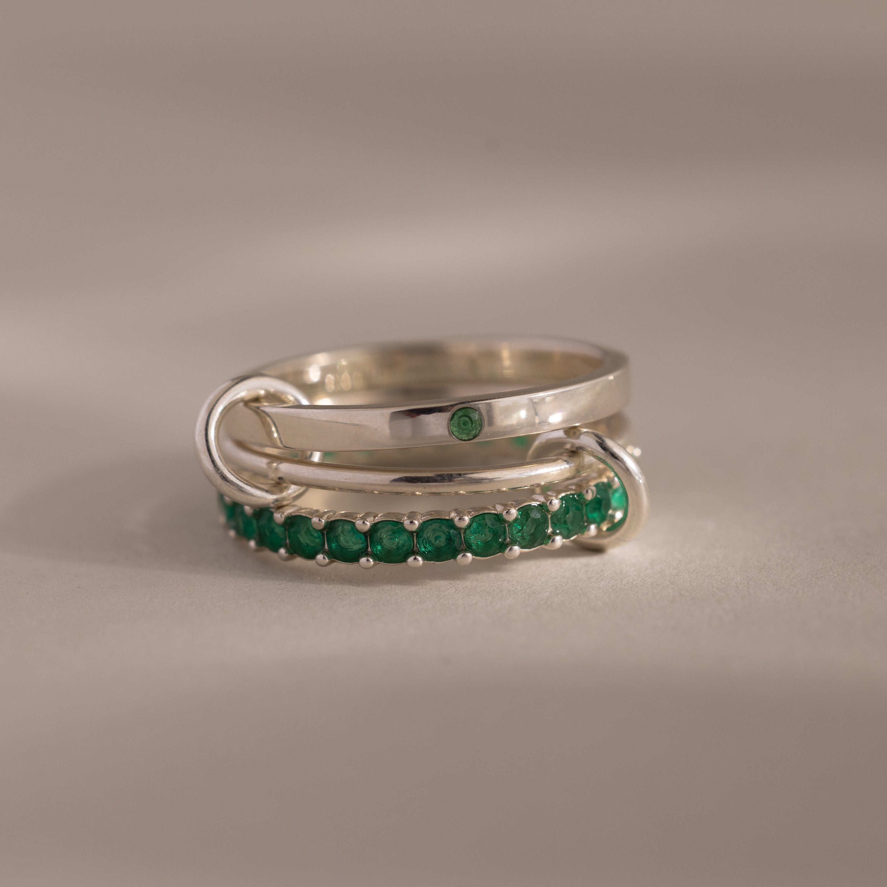 The Triple Linked Emerald Ring Set features three intertwined silver bands—one plain, one accented with a single green gem, and one lined with radiant green stones—symbolizing eternal connection in an elegant design.