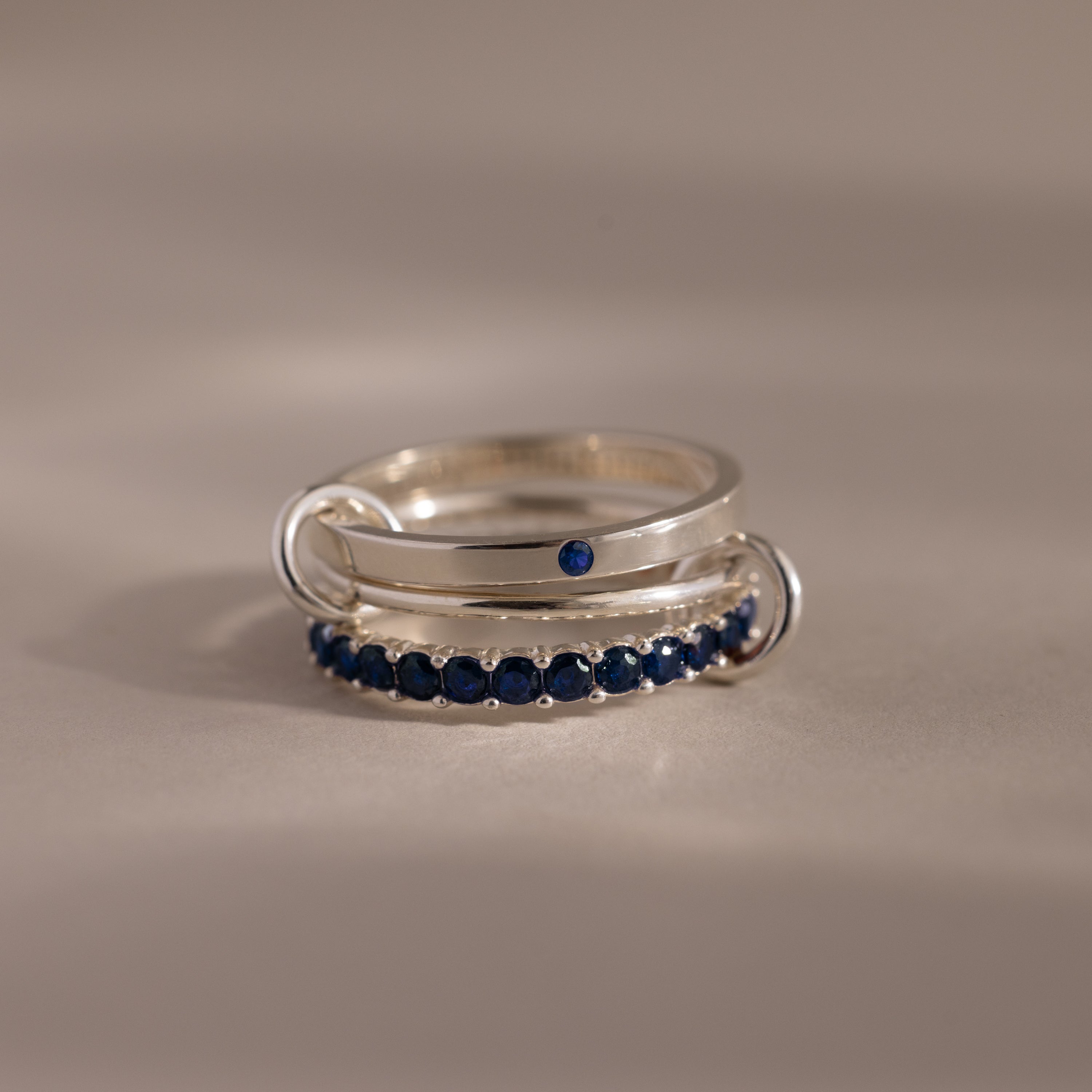 The Triple Linked Sapphire Ring Set features elegant silver bands adorned with a row of small blue gemstones and a single blue stone, shining beautifully on any neutral background.
