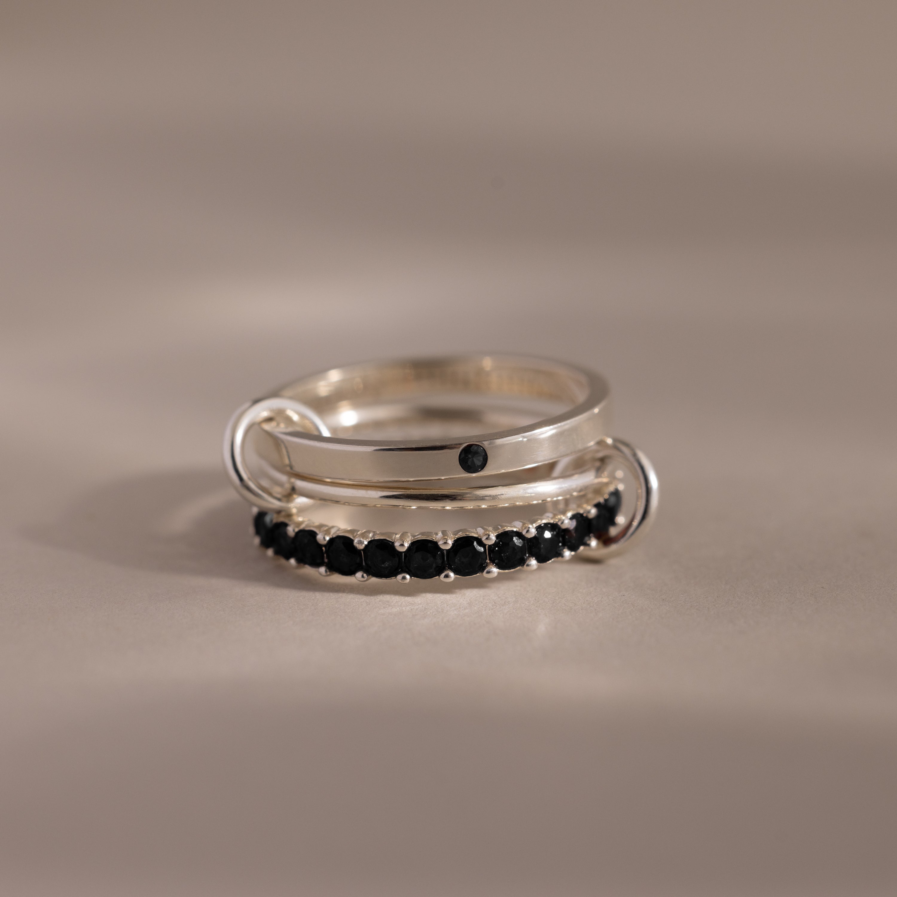The Triple Linked Onyx Ring Set, featuring a plain band with a black stone and an eternity band adorned with multiple stones, rests elegantly on a neutral surface.