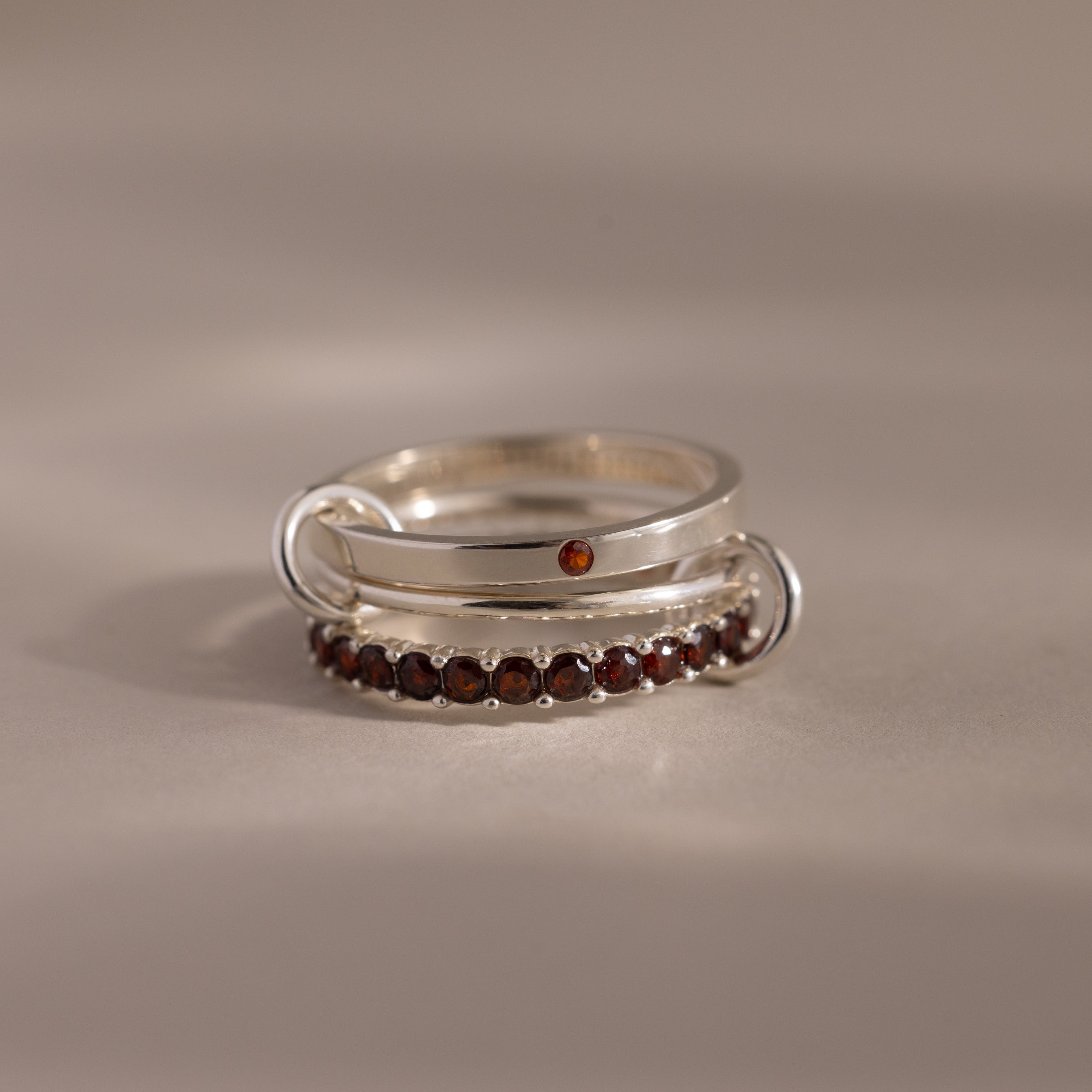 The Triple Linked Garnet Ring Set features three silver stacking rings, including an eternity band set with small red garnet stones, displayed on a beige surface.