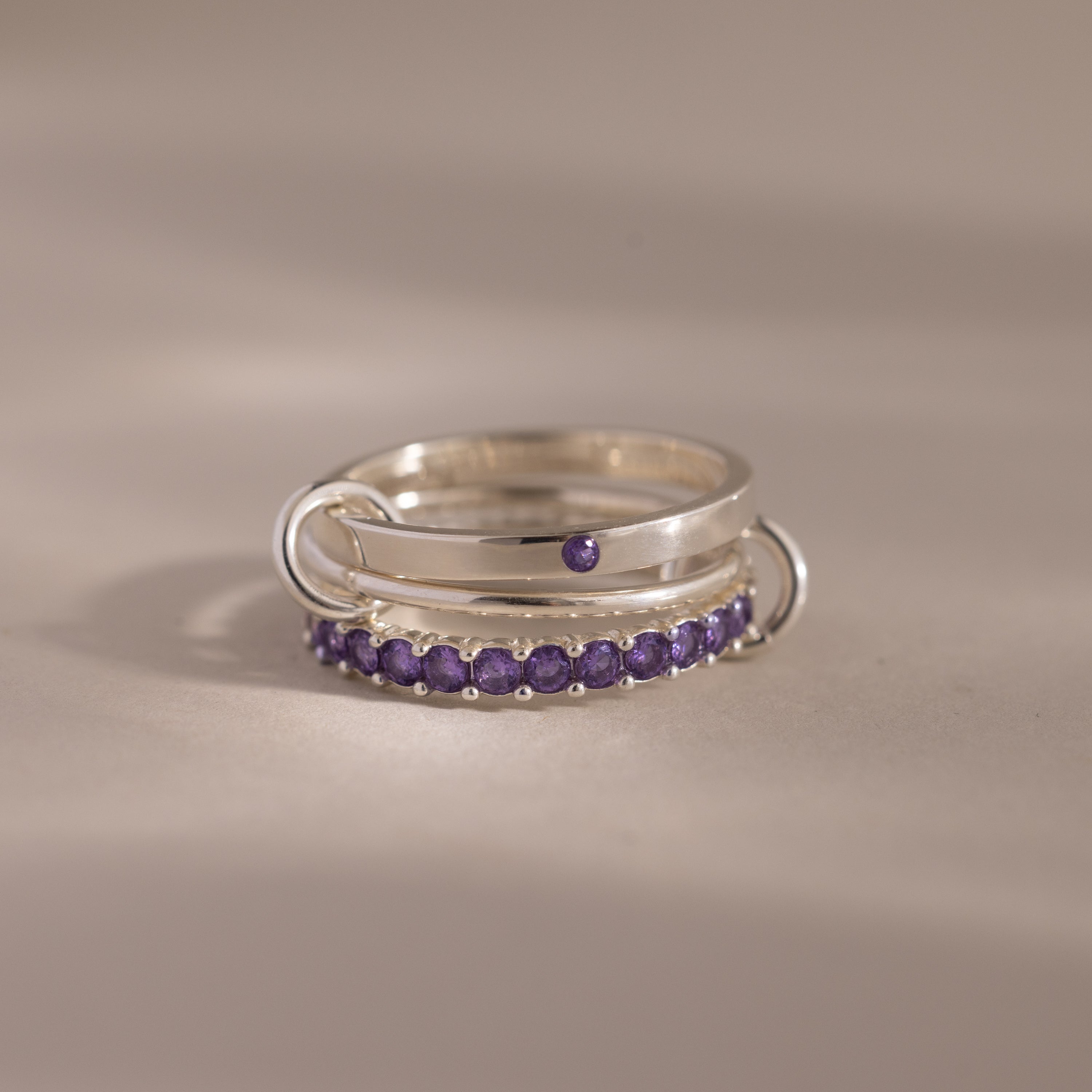 The Triple Linked Amethyst Ring Set features silver bands adorned with rows of small purple gemstones, displayed on a light surface.