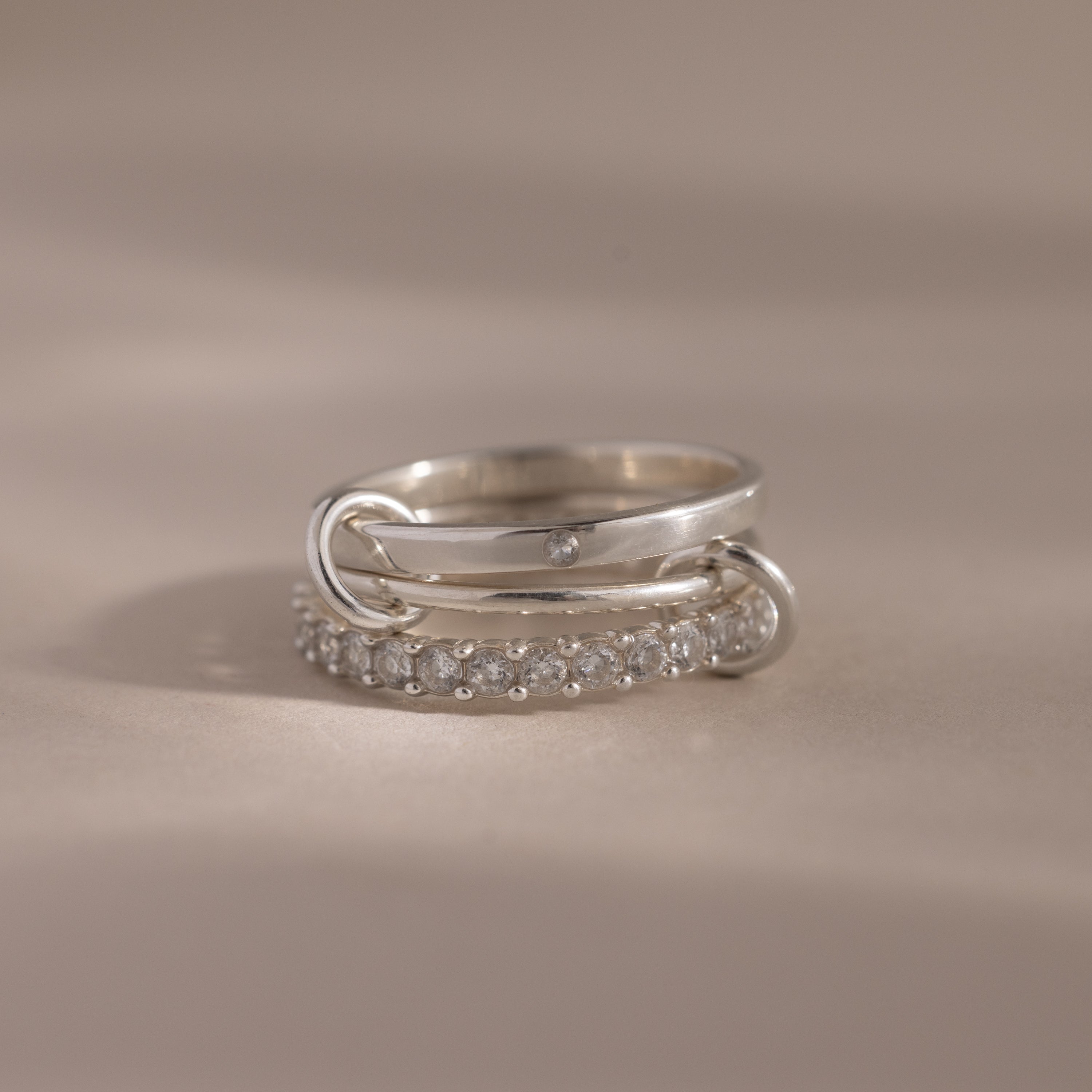 The Triple Linked Diamond Ring Set features a silver triple linked birthstone ring with a diamond and an eternity birthstone band adorned with small diamonds, displayed on a beige surface.