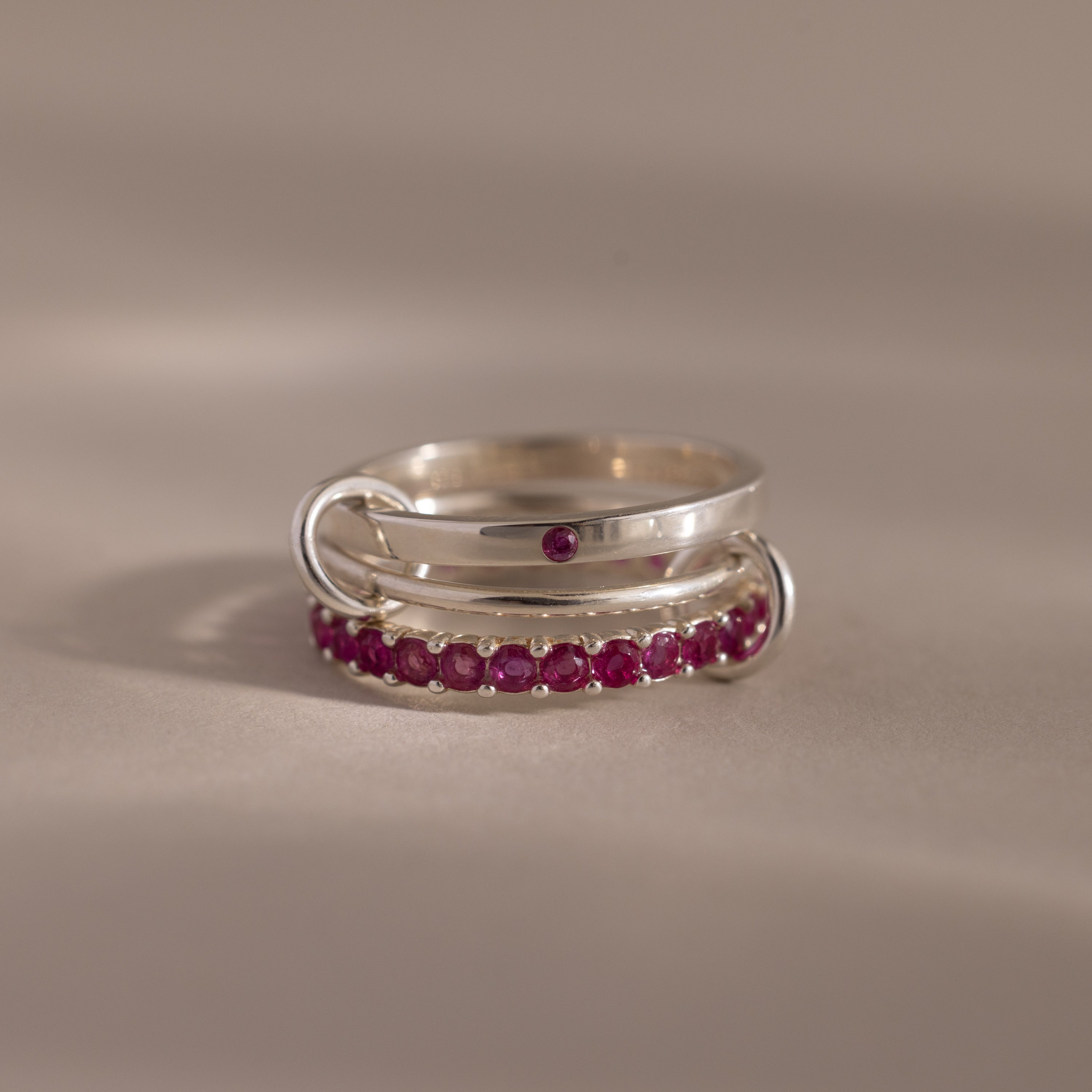 The Triple Linked Ruby Ring Set features a silver eternity band adorned with small red gemstones and one larger ruby, elegantly displayed on a neutral surface.