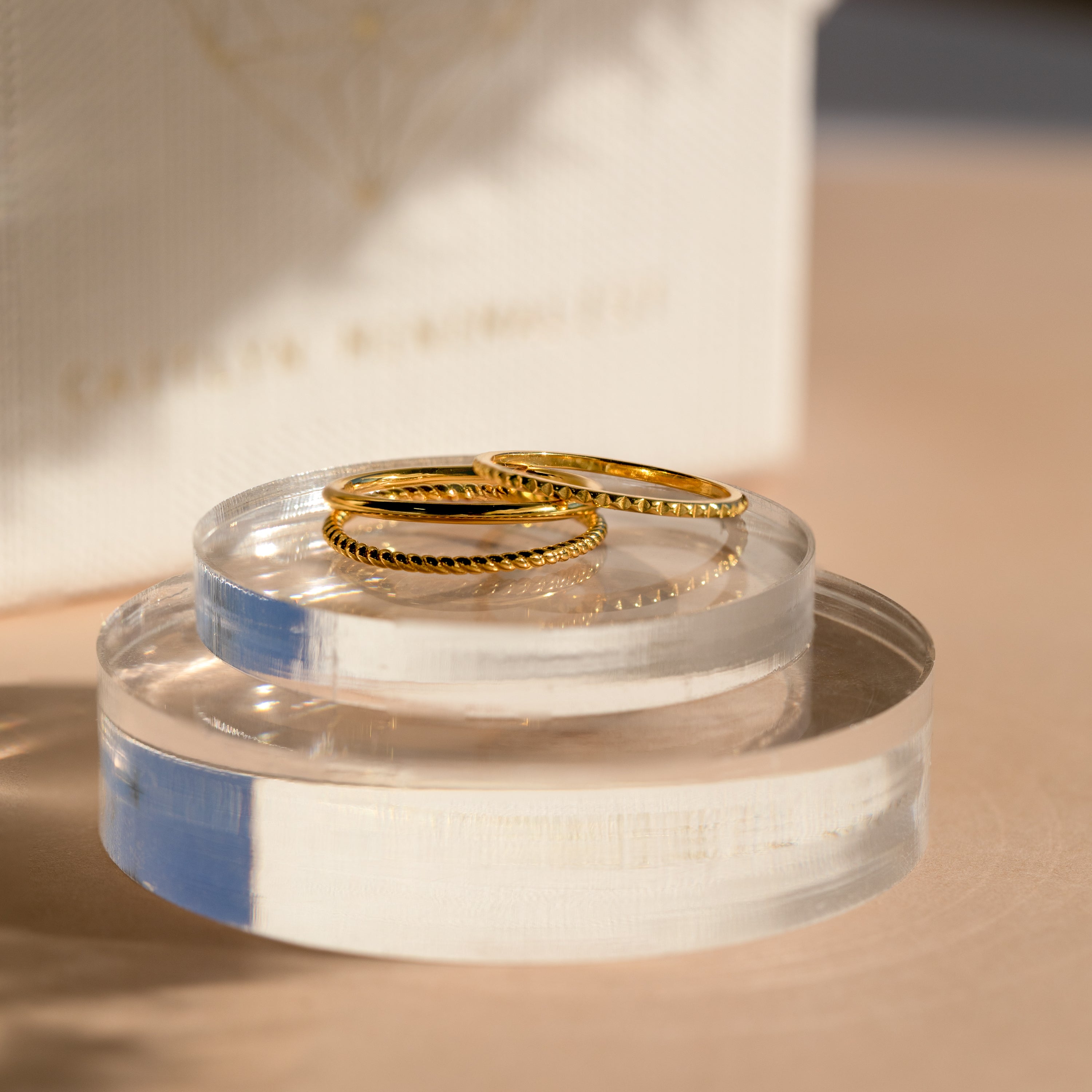 Two Thin Stacking Rings from our 18K Gold Set rest on clear acrylic stands in soft, natural light—a stylish ring stack and perfect Christmas gift.