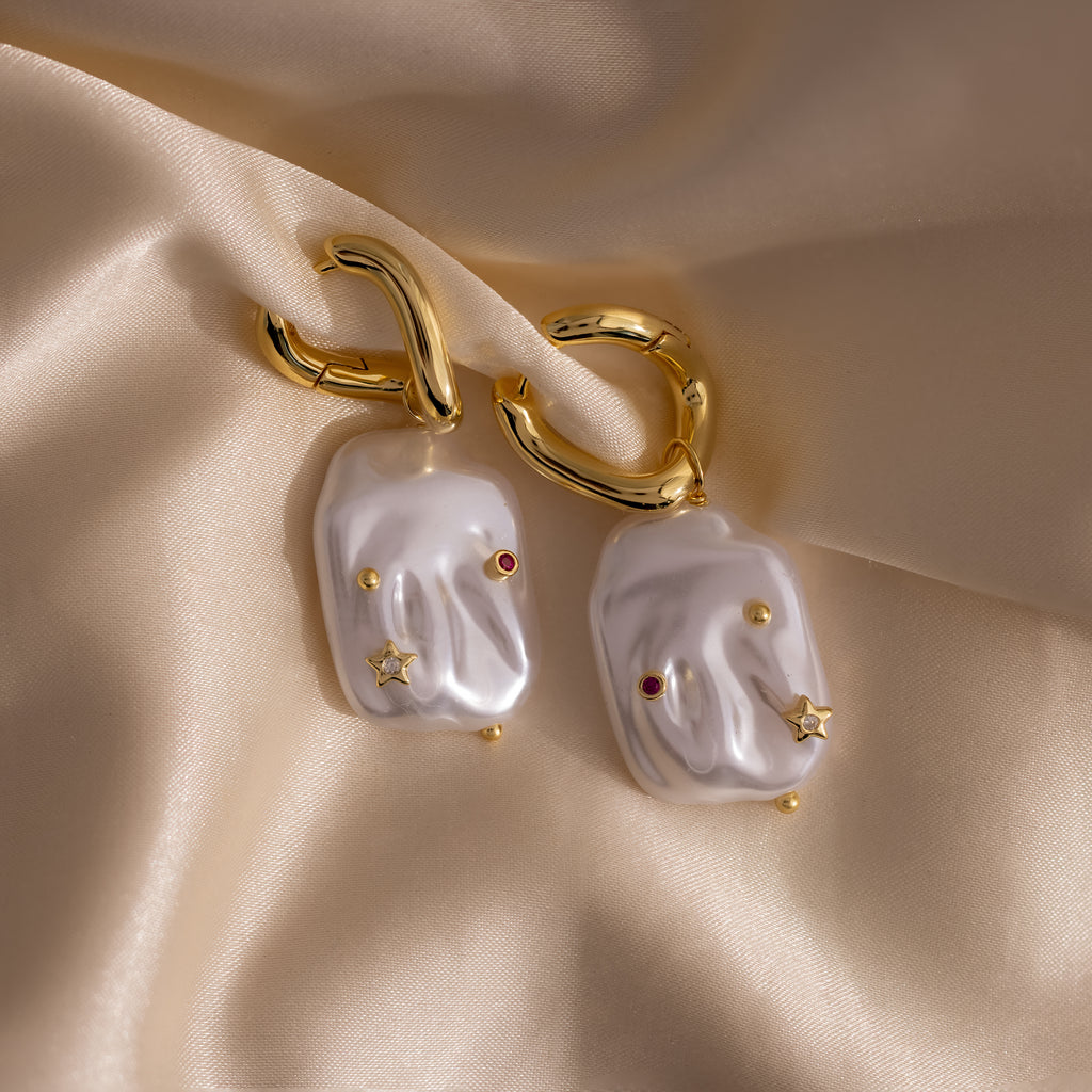 Celestial Baroque Pearl Charm Hoops featuring gold hoops with irregular baroque pearl drops, accented by star and gemstone charms, displayed on beige satin fabric.