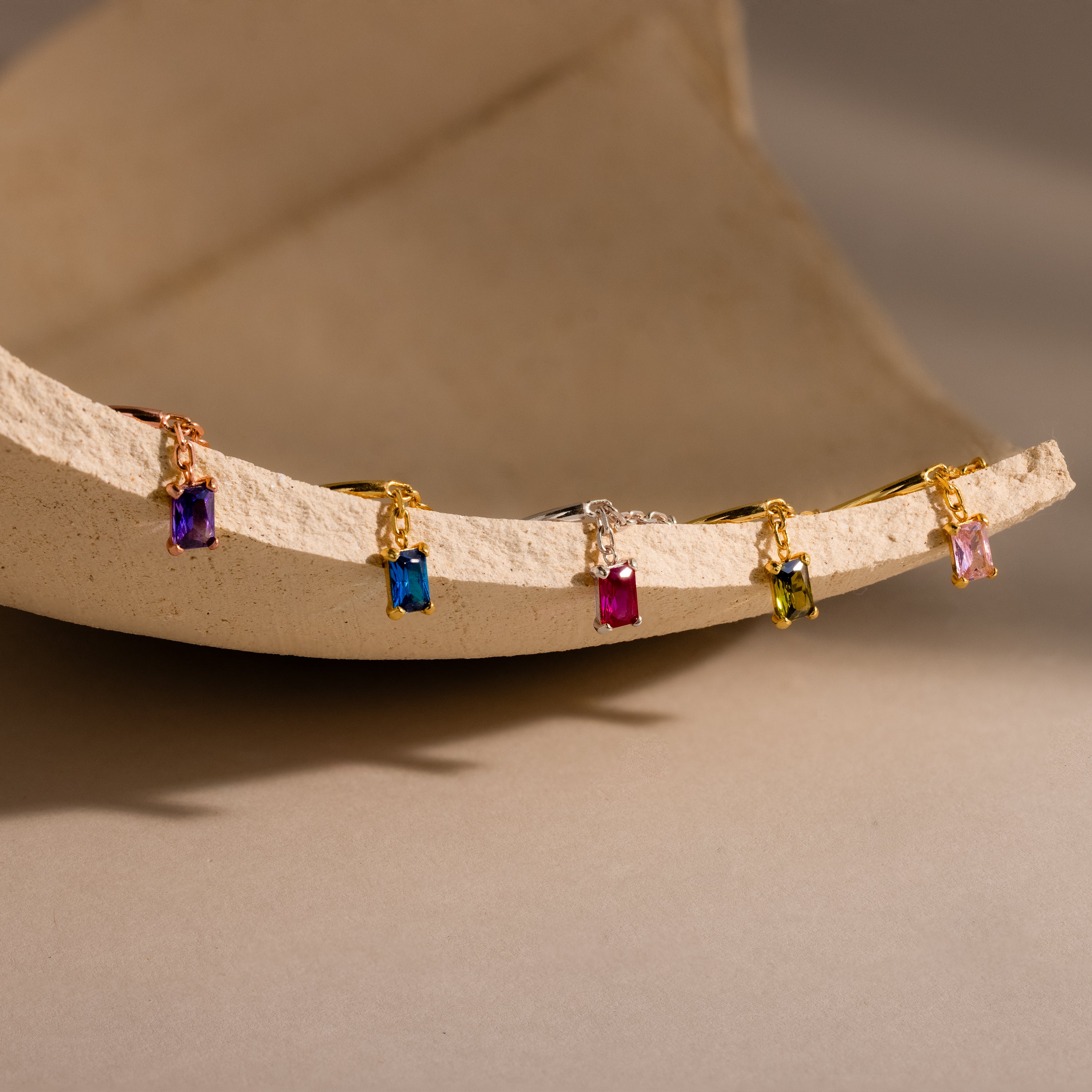 The Birthstone Drop Chain Ring, a dazzling gold ring with colorful gemstones, is displayed alongside four other elegant gold rings on a textured, curved beige surface.