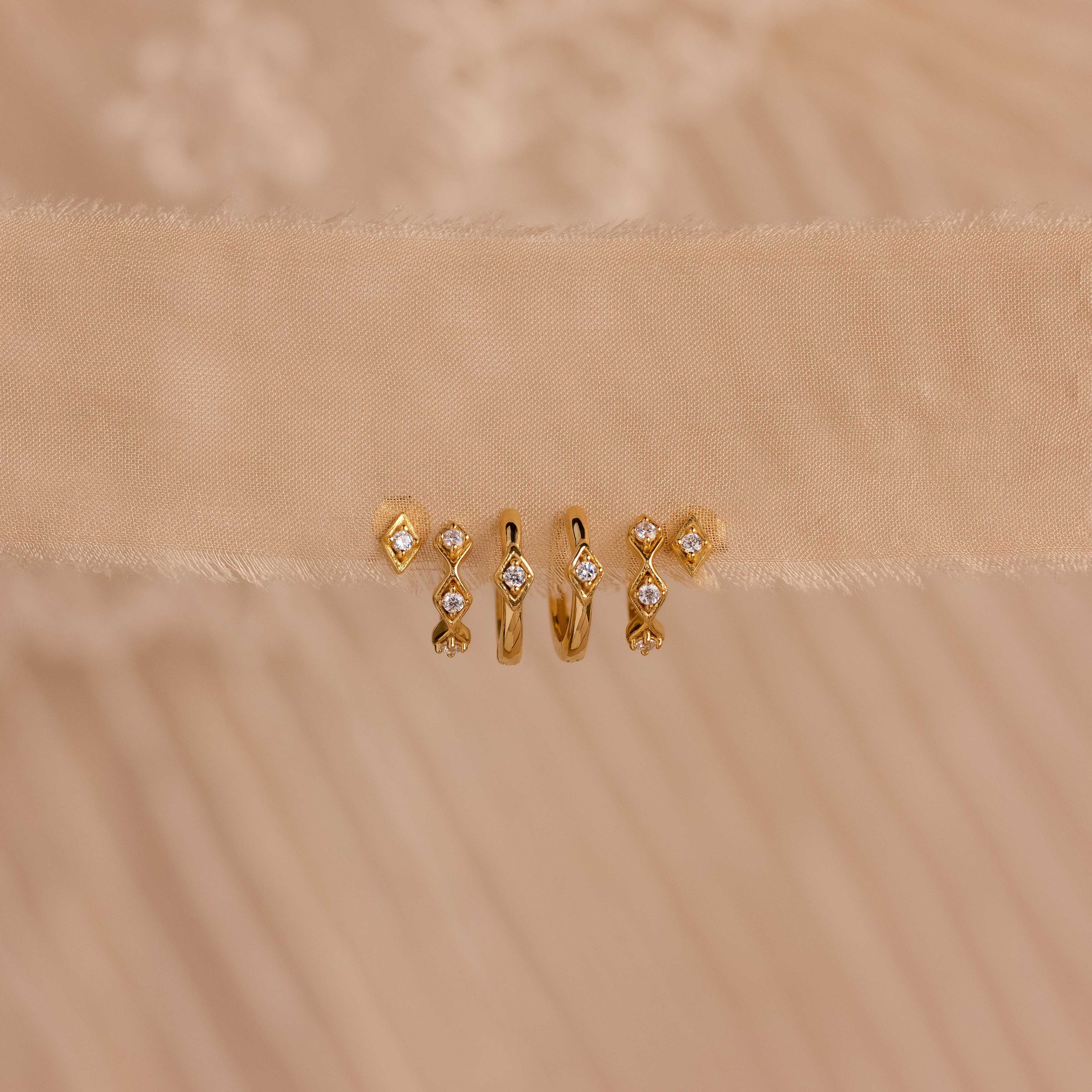 Five gold earrings, including the Ericka Diamond Studs with small diamonds, plus vintage-inspired and minimalist designs, are displayed on a beige textured fabric background.