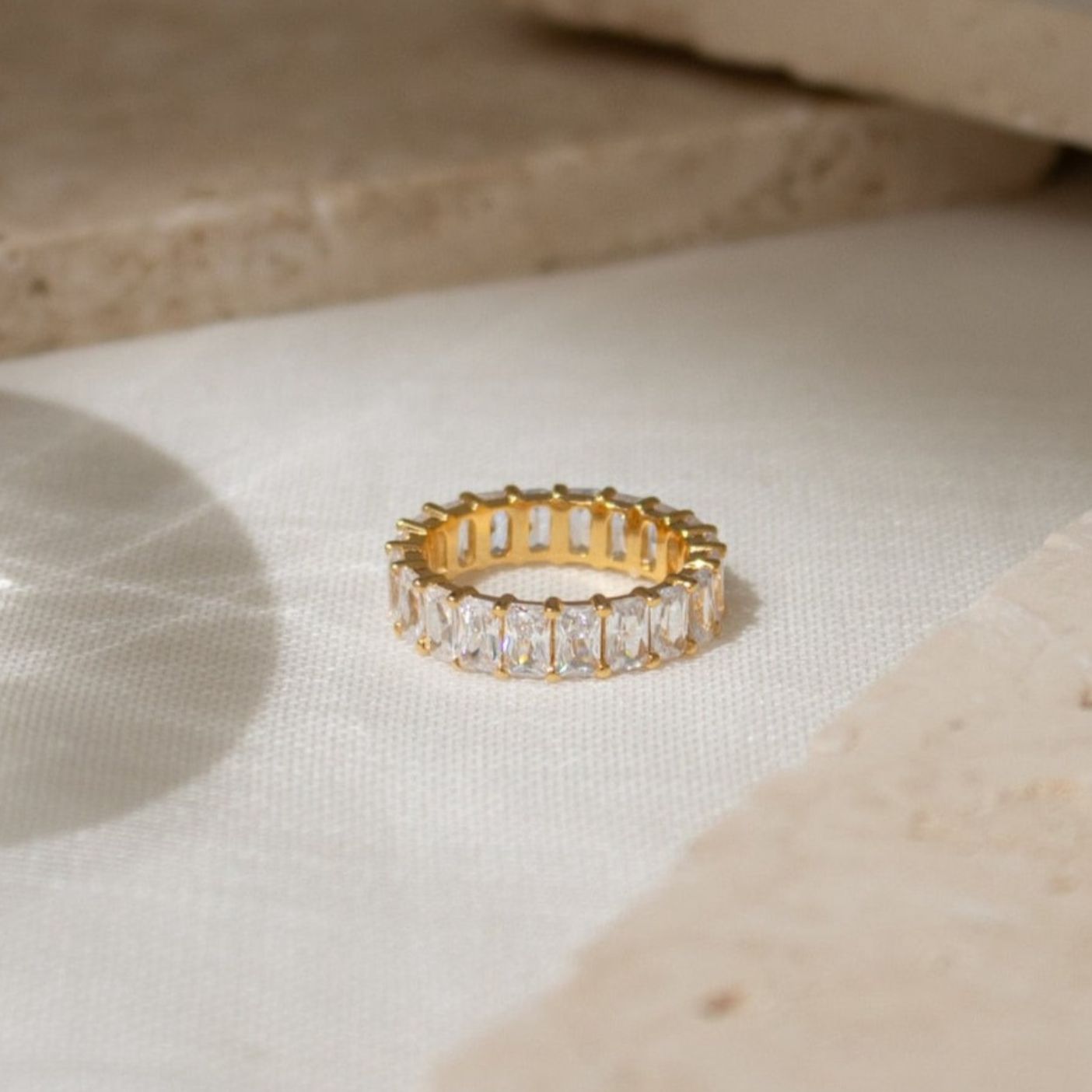 The Colette Eternity Ring, featuring rectangular-cut baguette diamonds in gold, is displayed on a light fabric surface beside beige stone slabs.
