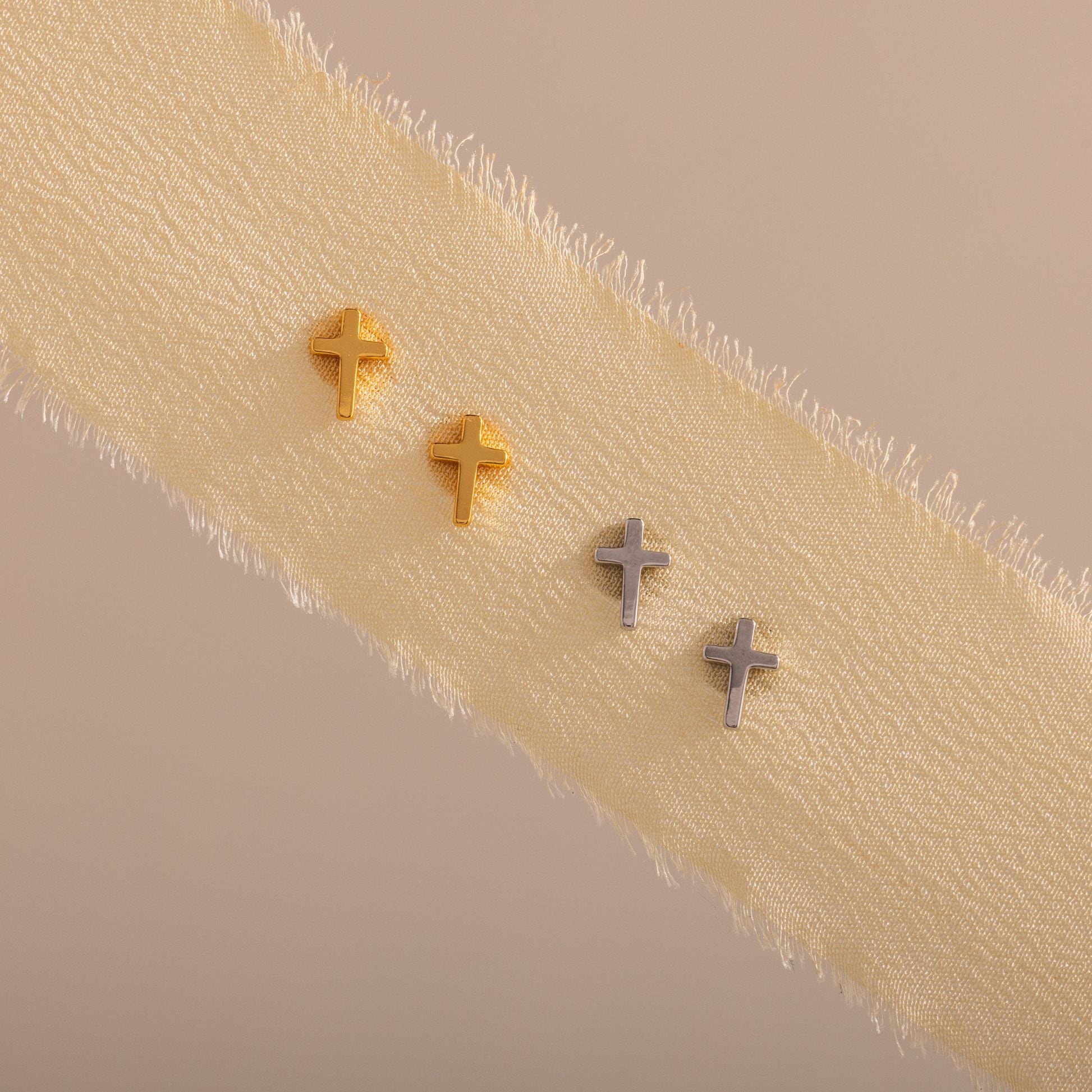 Gold and silver cross stud earrings displayed on a textured cream ribbon.