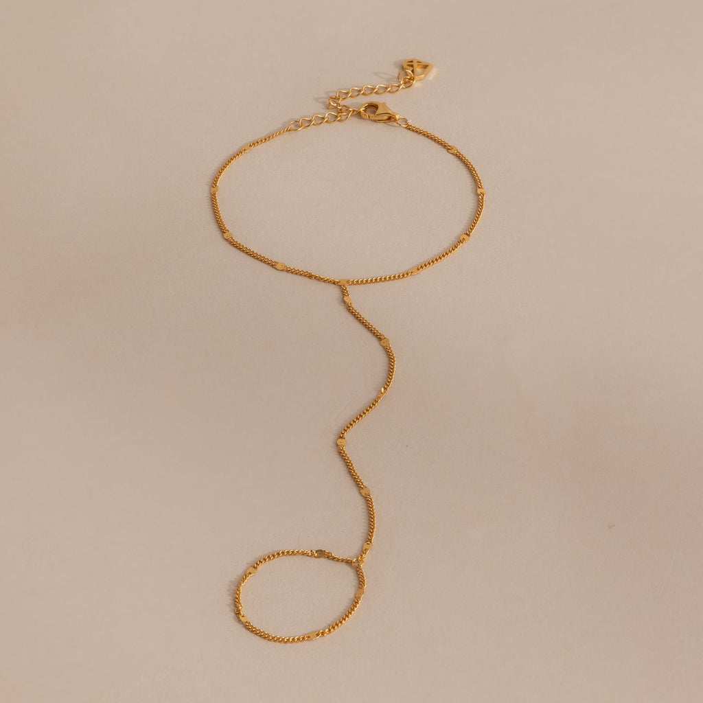Delicate curb hand chain bracelet in 18K gold resting on a neutral surface.