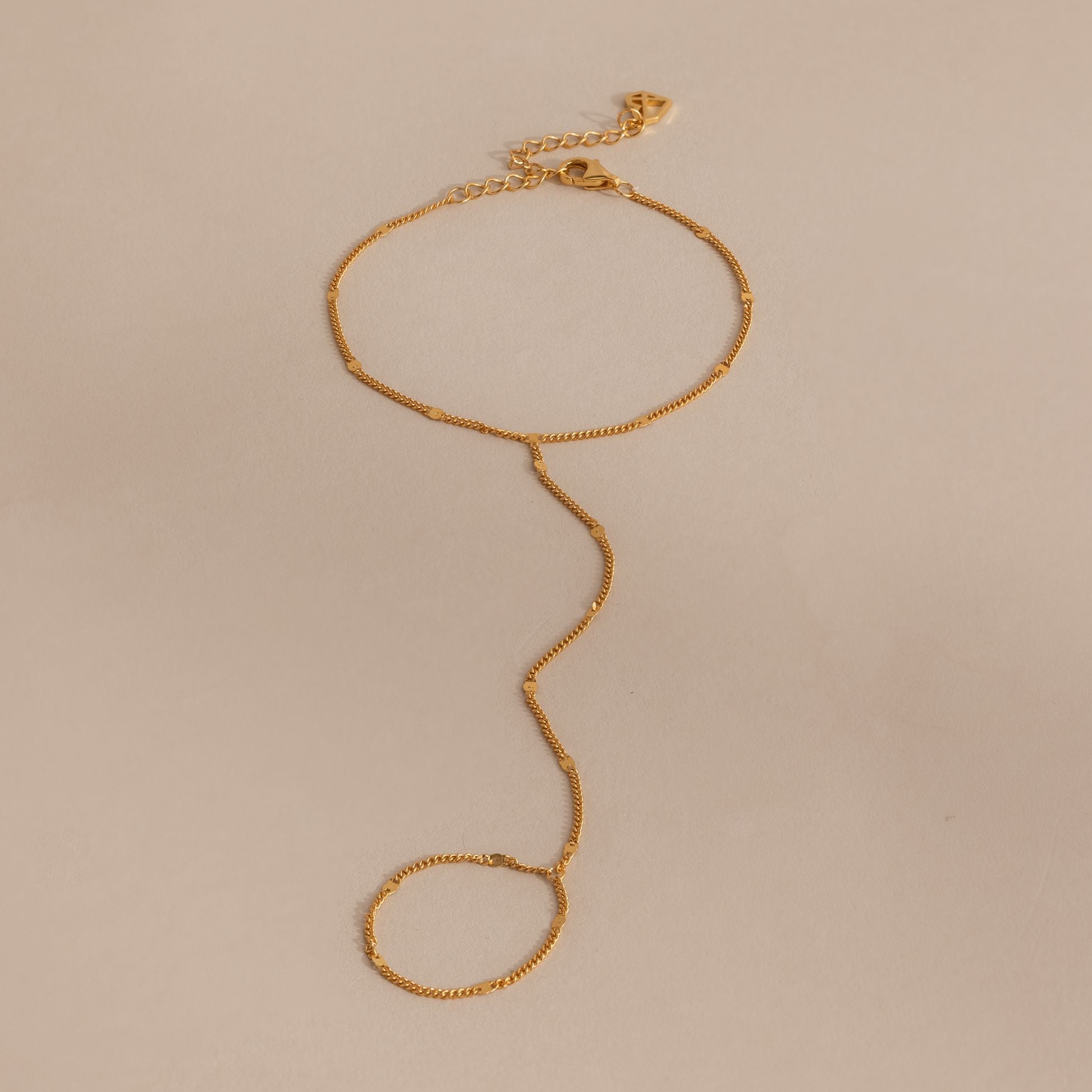 Delicate curb hand chain bracelet in 18K gold resting on a neutral surface.