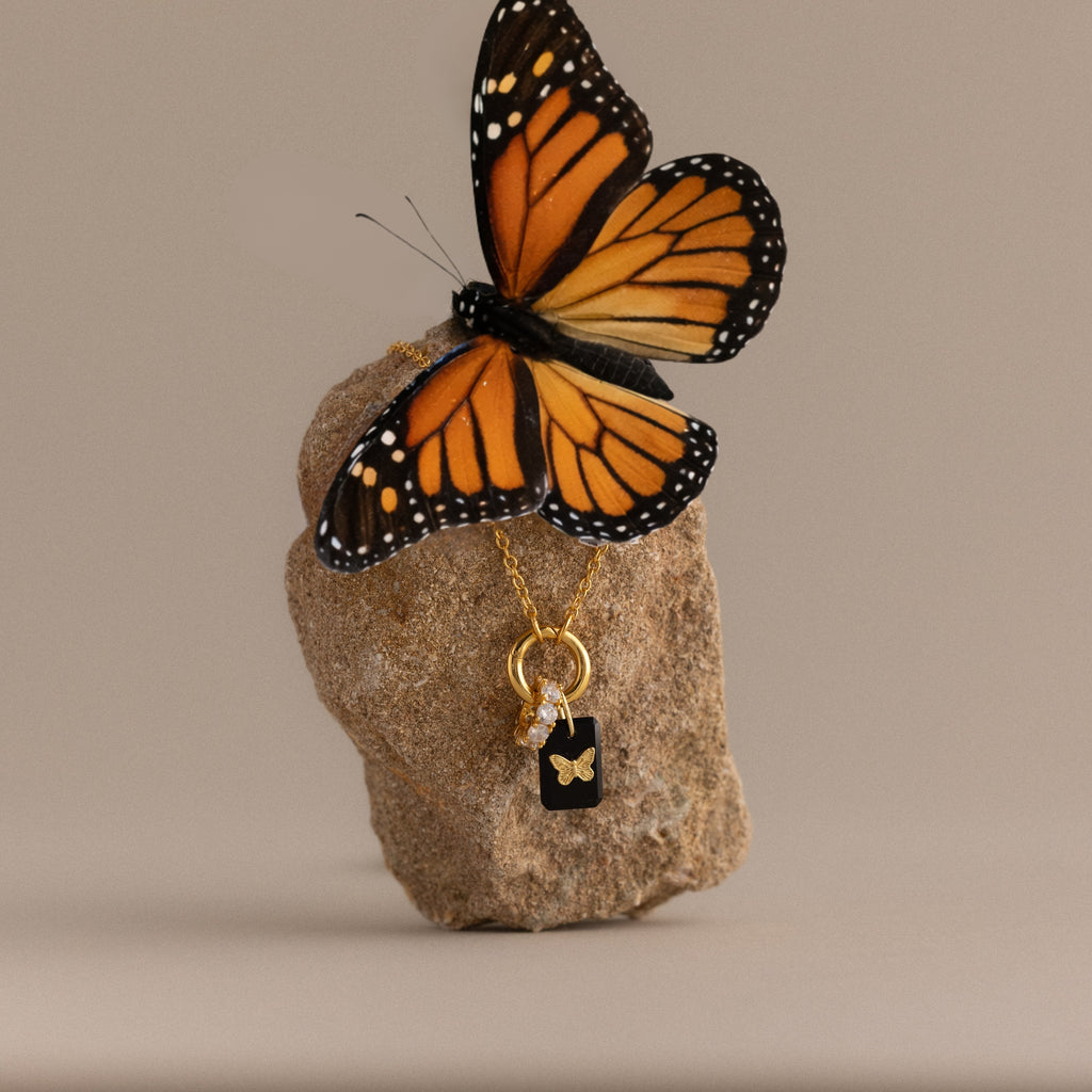 A monarch butterfly perched on a rock is adorned with the Onyx Butterfly Charm in 18K Gold, featuring a delicate butterfly pendant.