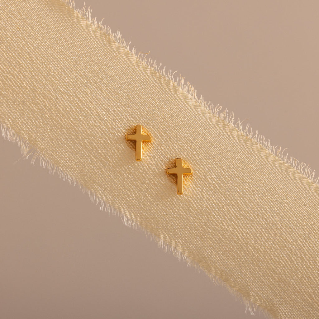 Dainty gold cross stud earrings displayed on a neutral ribbon fabric.