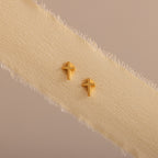 Dainty gold cross stud earrings displayed on a neutral ribbon fabric.