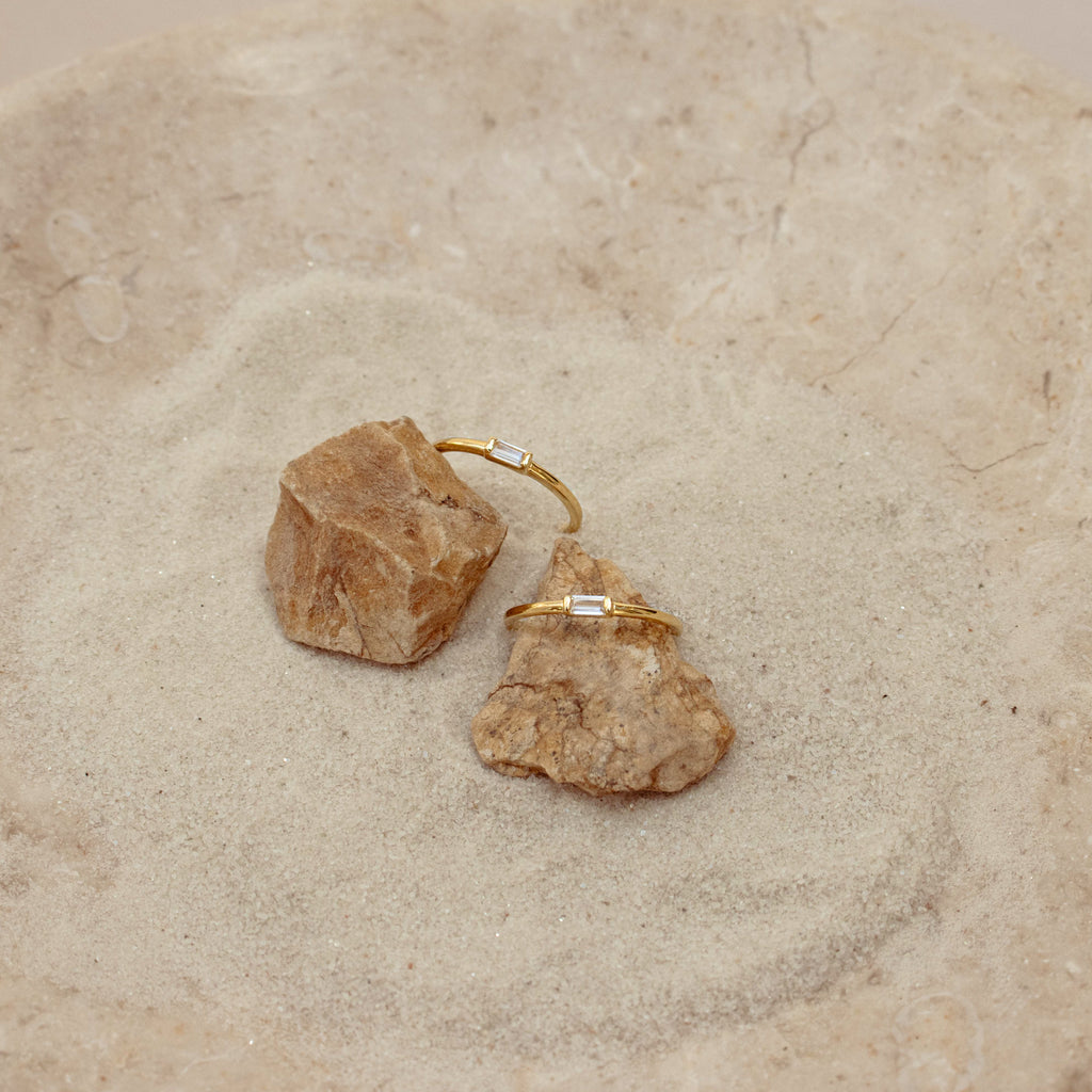 Two Baguette Diamond Rings with delicate baguette diamonds in an east-west bezel setting, displayed on rocks in a sand-filled dish.