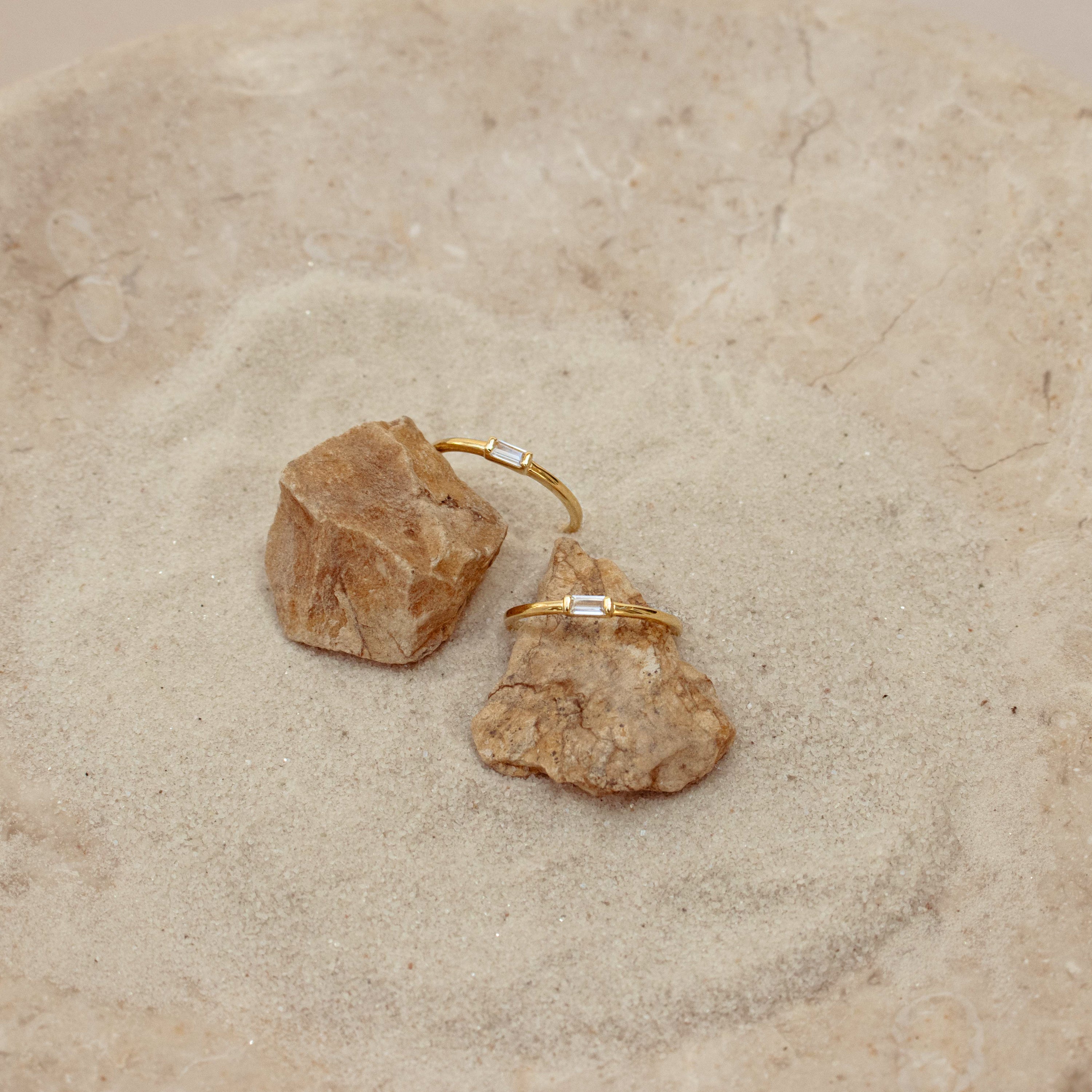 Two Baguette Diamond Rings with delicate baguette diamonds in an east-west bezel setting, displayed on rocks in a sand-filled dish.