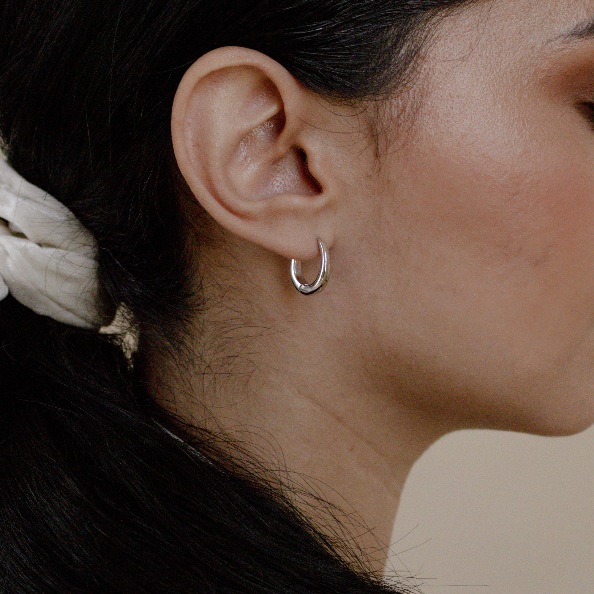 Close-up of a woman's ear adorned with Paris Oval Hoops in Sterling Silver—her dark hair tied back with a white scrunchie.