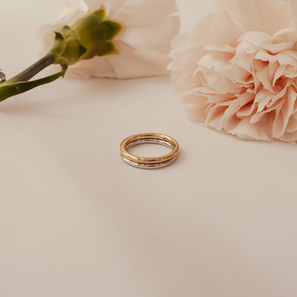 The Minimal Stacking Ring is a delicate gold wedding band, photographed on a white surface beside two pale pink carnations—ideal for minimalist bridal or everyday stacking styles.