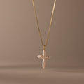 The Baroque Pearl Cross Necklace in 18K Gold features a white freshwater pearl cross pendant on a gold chain, elegantly displayed against a neutral beige background.
