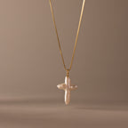 The Baroque Pearl Cross Necklace in 18K Gold features a white freshwater pearl cross pendant on a gold chain, elegantly displayed against a neutral beige background.