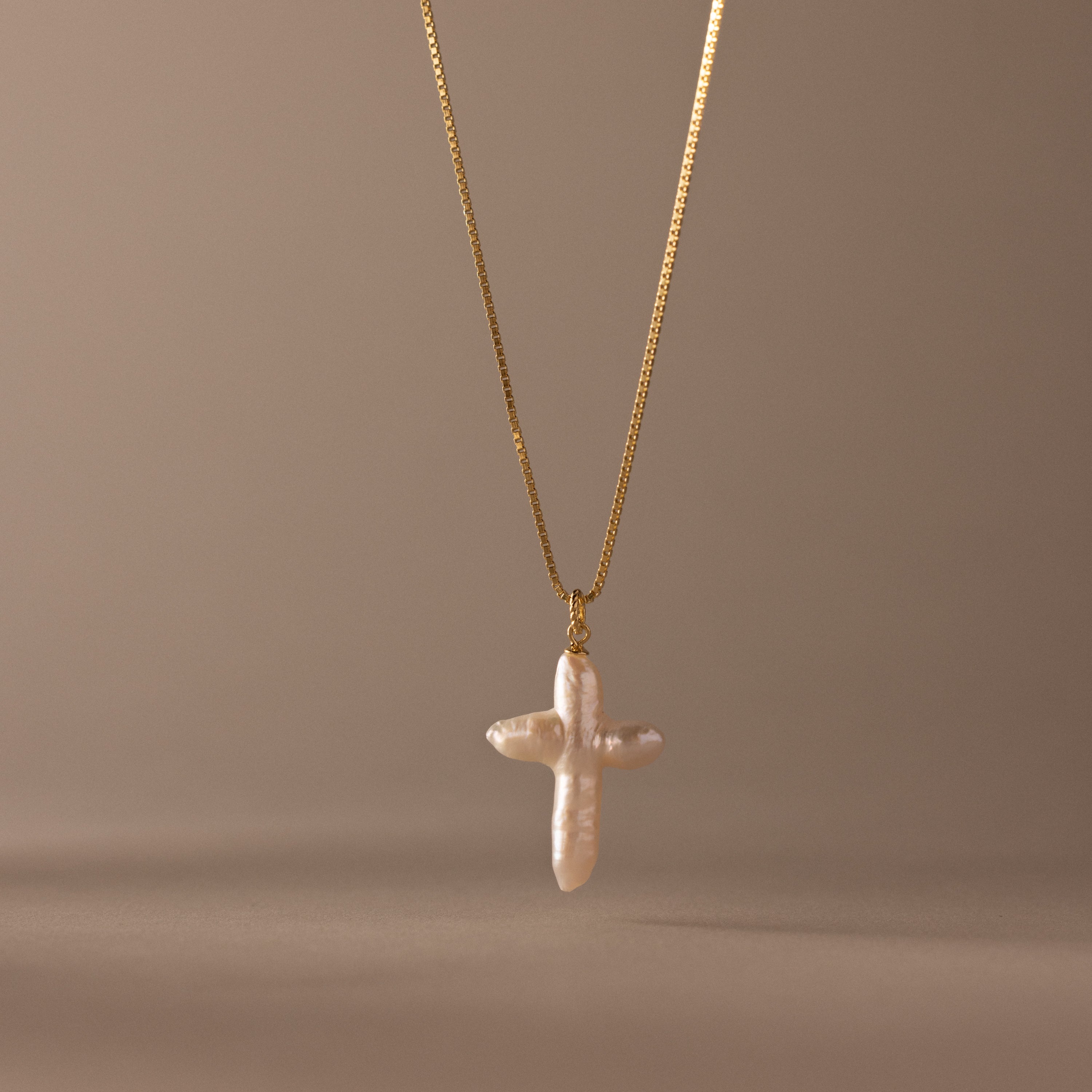 The Baroque Pearl Cross Necklace in 18K Gold features a white freshwater pearl cross pendant on a gold chain, elegantly displayed against a neutral beige background.
