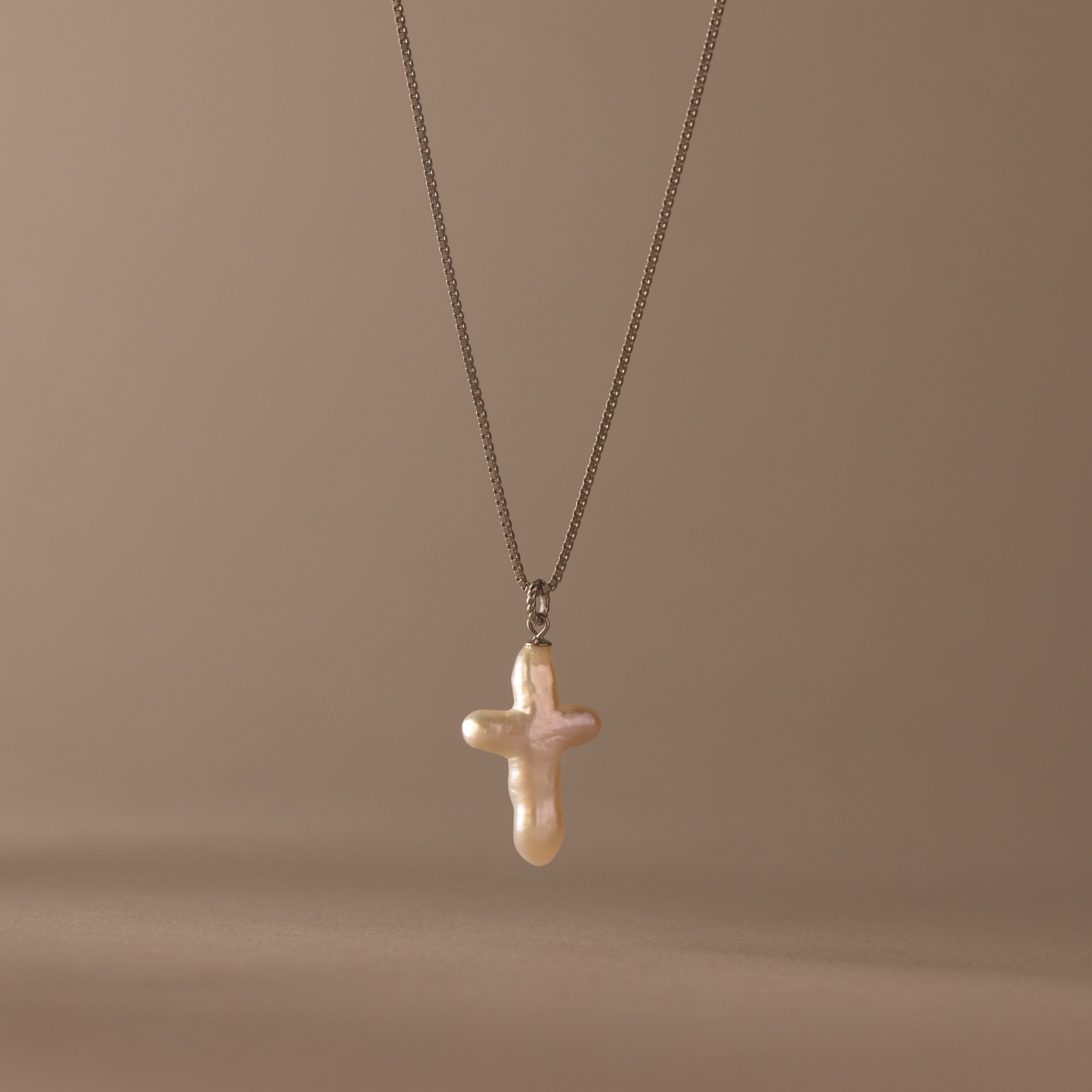 The Baroque Pearl Cross Necklace in Silver showcases a cross-shaped baroque pearl pendant on a minimalist chain, displayed against a soft beige background.