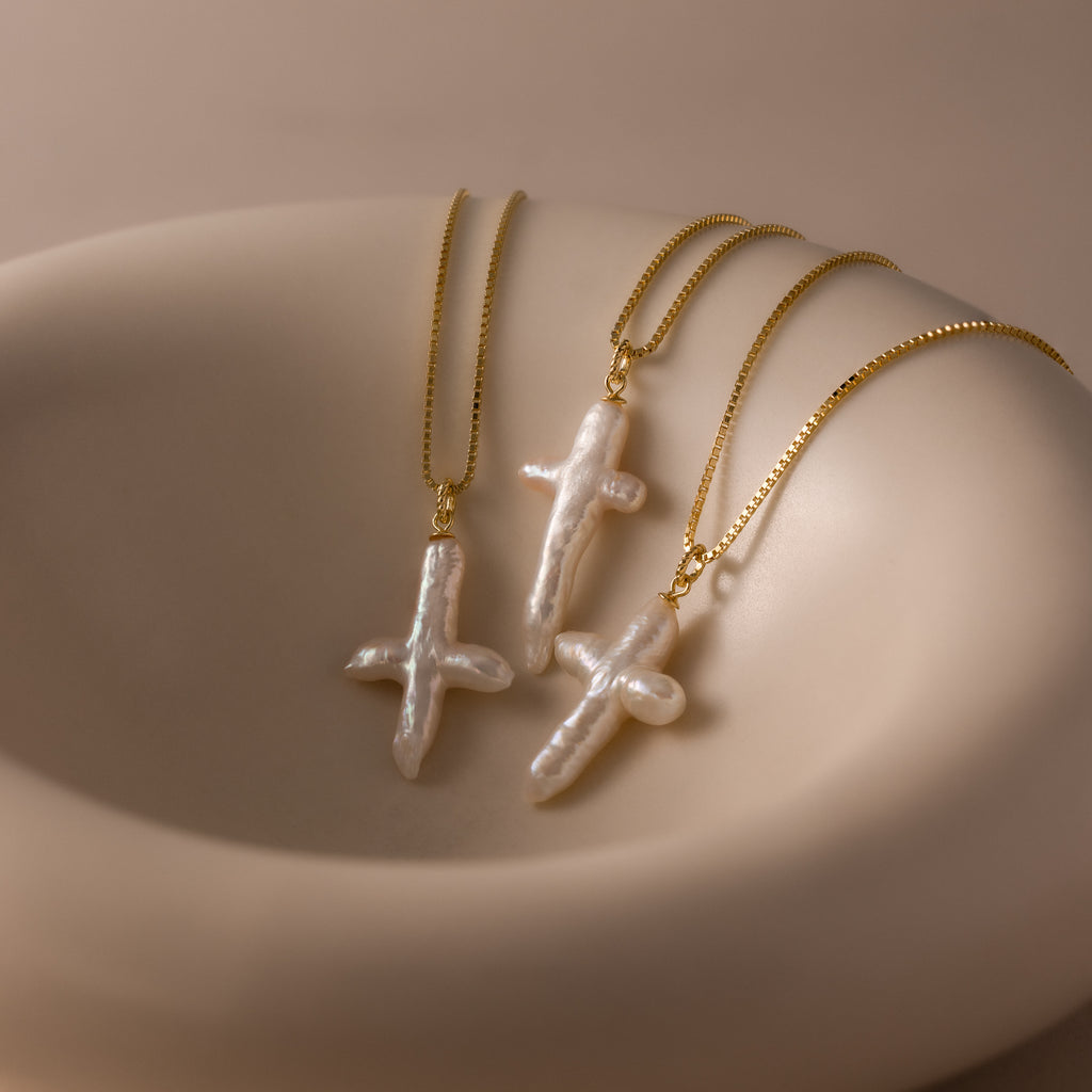 Three gold chain necklaces, including the Baroque Pearl Cross Necklace in 18K Gold and a freshwater pearl necklace, are displayed on a smooth beige surface.