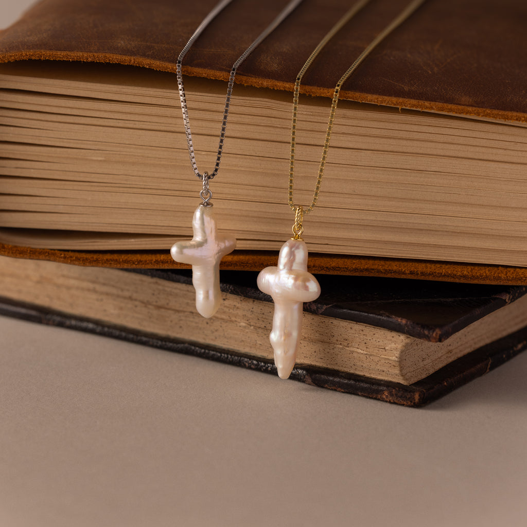 Two Baroque Pearl Cross Necklaces—one with a silver chain, one with a gold chain—are displayed on stacked books, offering a perfect minimalist look.