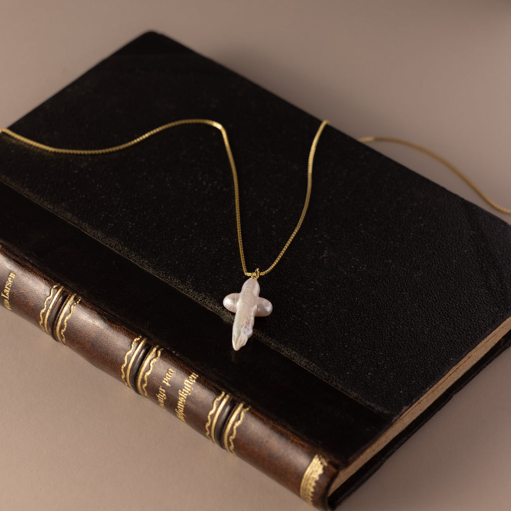 The Baroque Pearl Cross Necklace in 18K Gold rests on stacked vintage books, its luminous freshwater pearls and elegant gold chain bringing timeless sophistication to the scene.