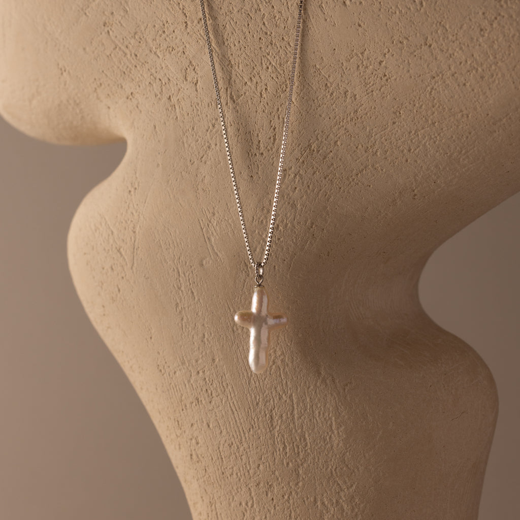 The Baroque Pearl Cross Necklace showcases a pearl cross pendant on a silver chain, elegantly displayed on a beige textured mannequin bust.