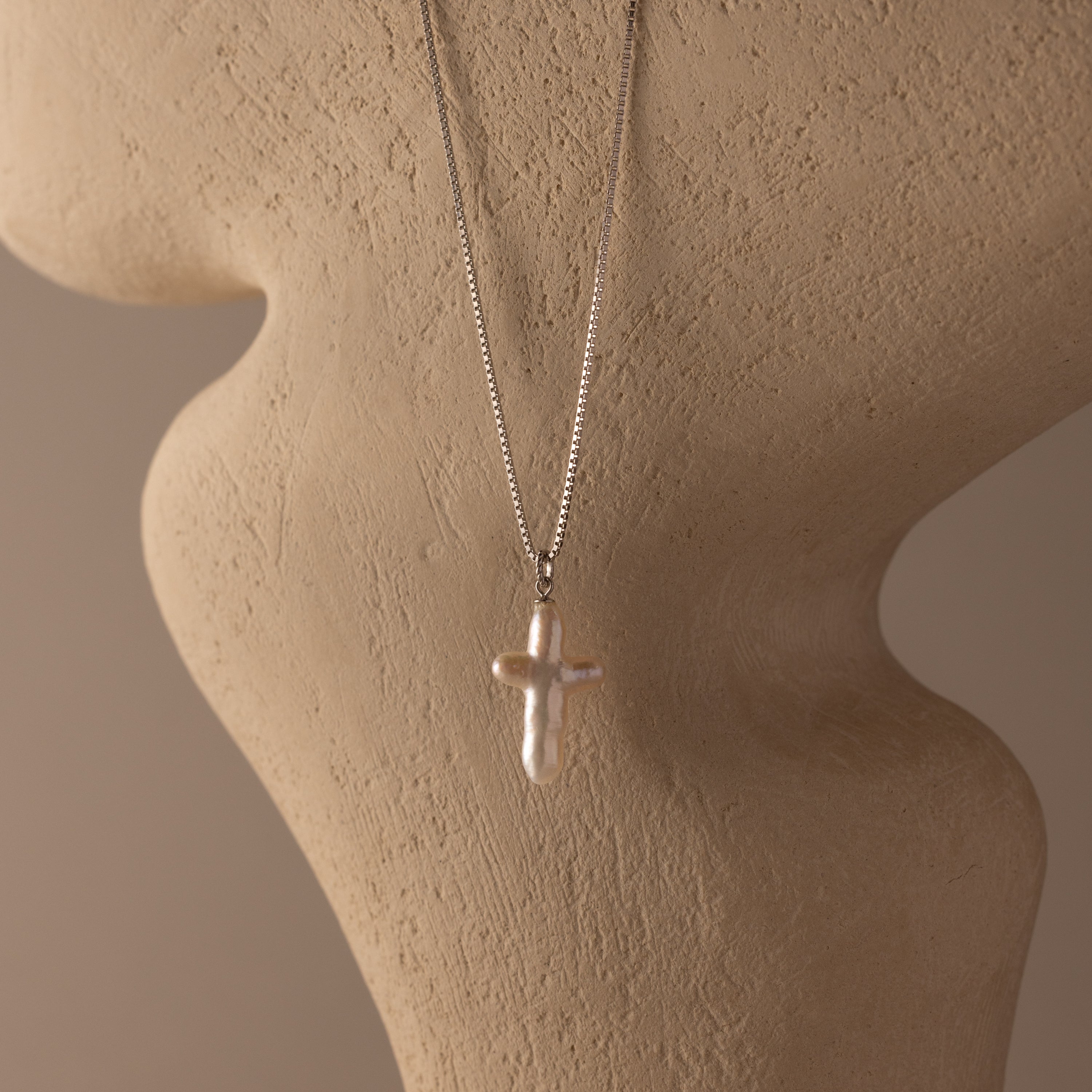 The Baroque Pearl Cross Necklace showcases a pearl cross pendant on a silver chain, elegantly displayed on a beige textured mannequin bust.
