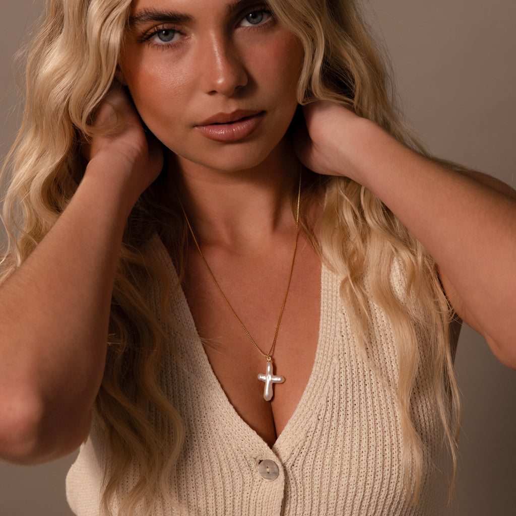 A woman with long blonde hair wears a cream knit top and the Baroque Pearl Cross Necklace, which features freshwater pearls and a gold chain for a minimalist look.