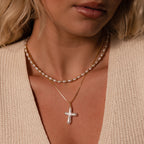 A woman wears a freshwater pearl necklace and the Baroque Pearl Cross Necklace in 18K Gold, styled with a minimalist gold chain and a beige knit top.