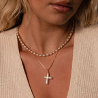 Woman wearing a beige knit top, a pearl necklace, and the Baroque Pearl Cross Necklace, which features a gold chain and pearl cross pendant.